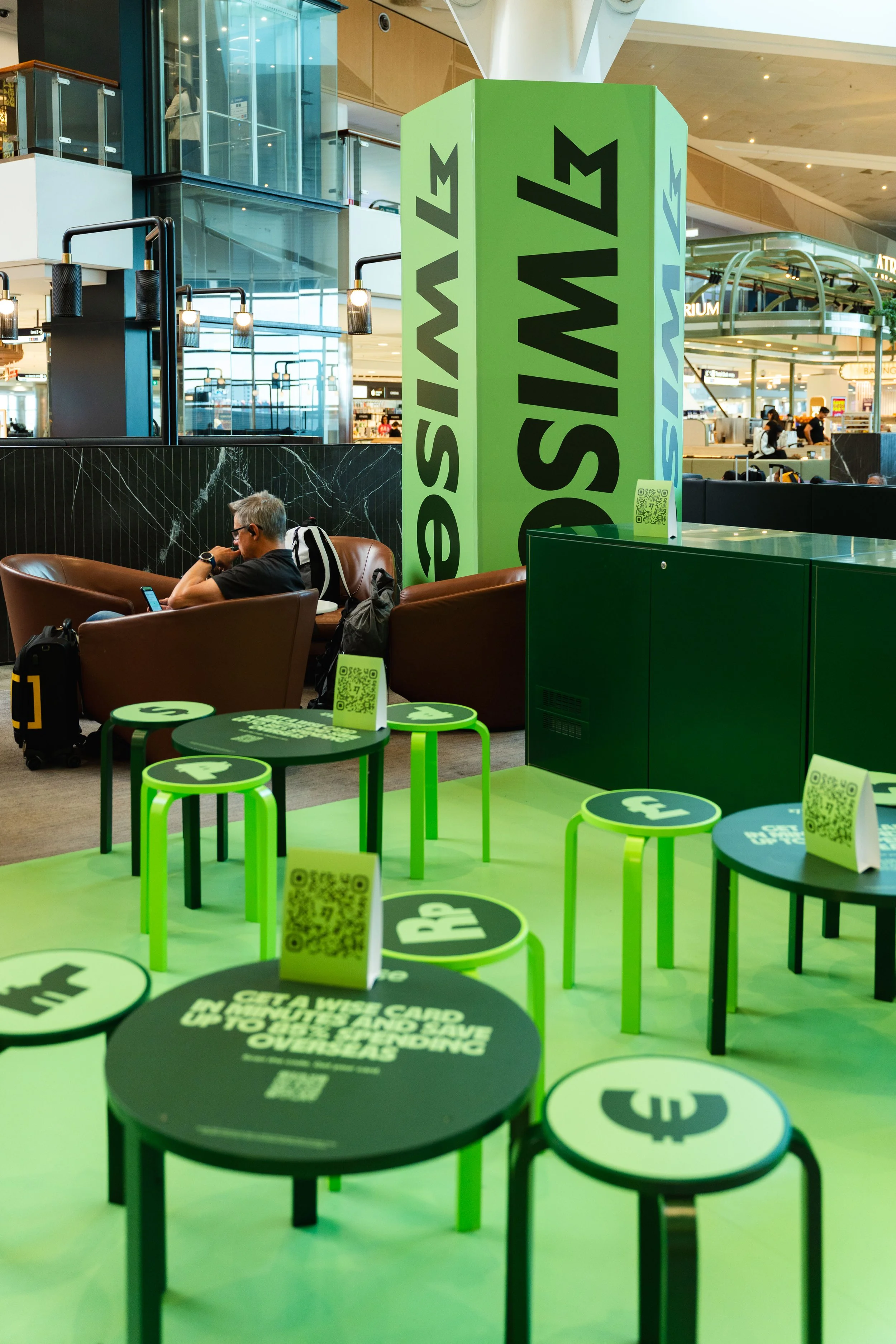Airport seating area with a large green sign reading 'SWISS' and several smaller tables and chairs with QR codes and the Swiss cross in a public space.