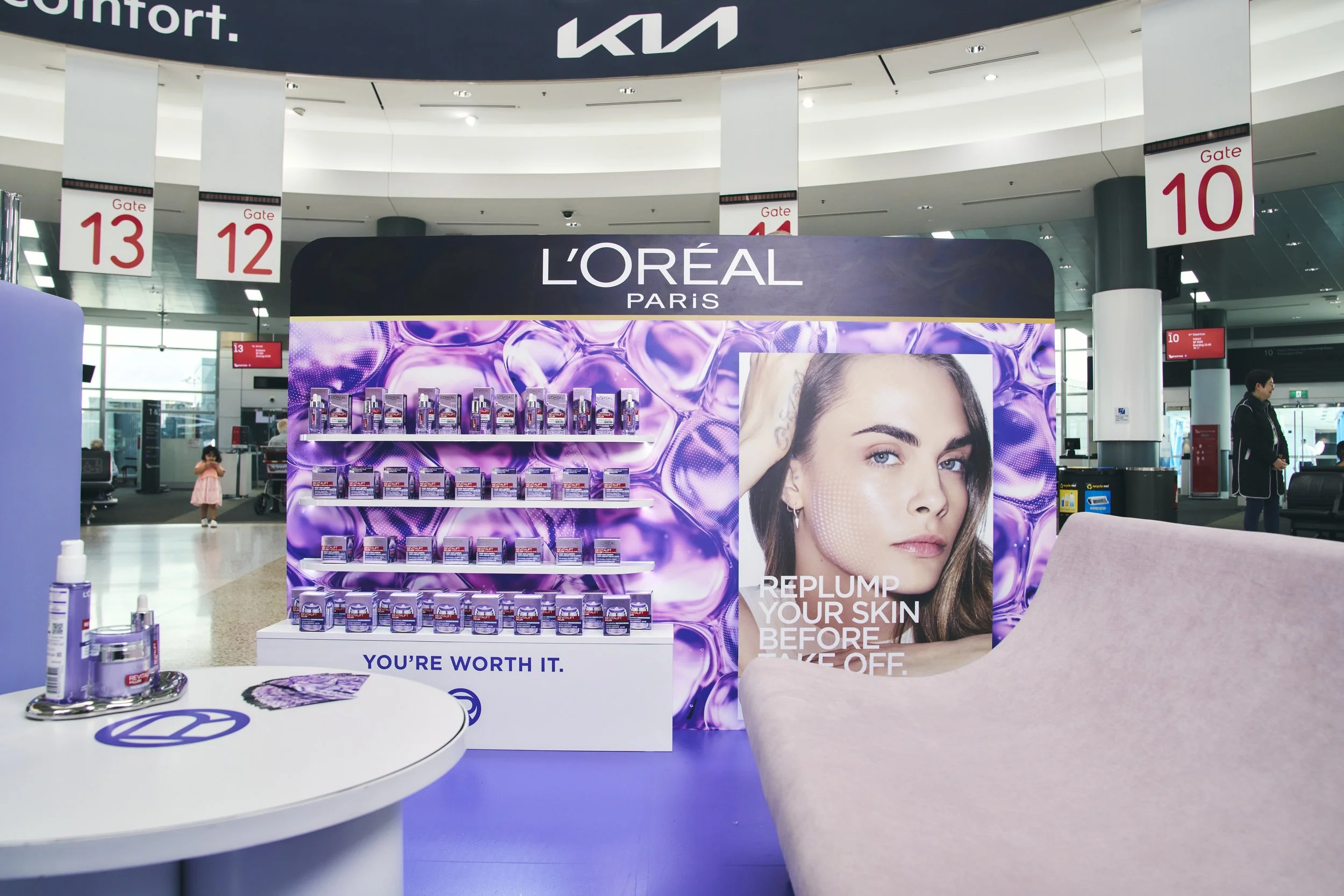 KIA and L'Oréal Paris promotional booth in an airport, with a large poster of a woman promoting skincare products, and shelves displaying L'Oréal products.
