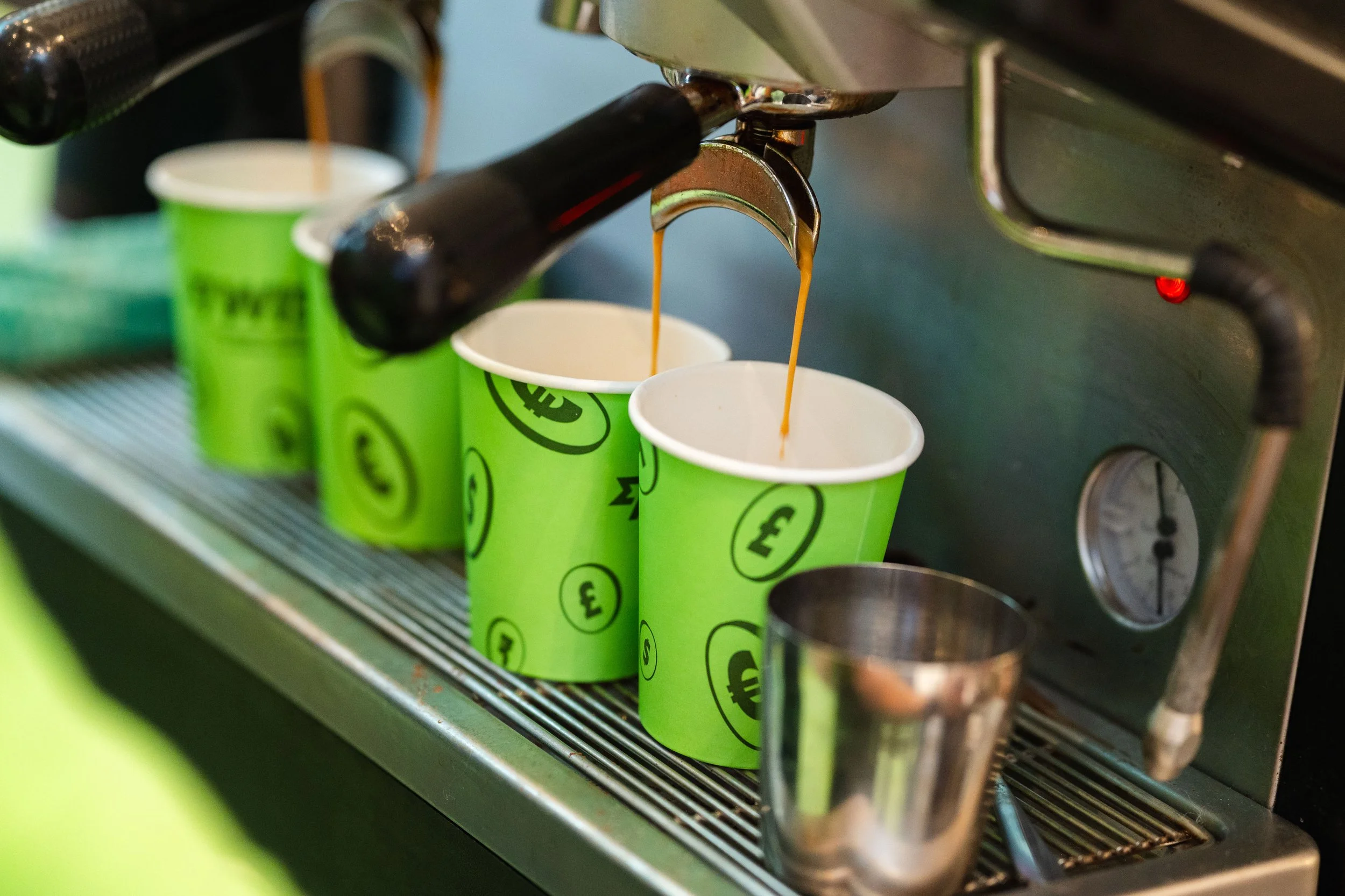 Espresso machine pouring coffee into green cups with currency symbols, with a metal shot glass nearby.