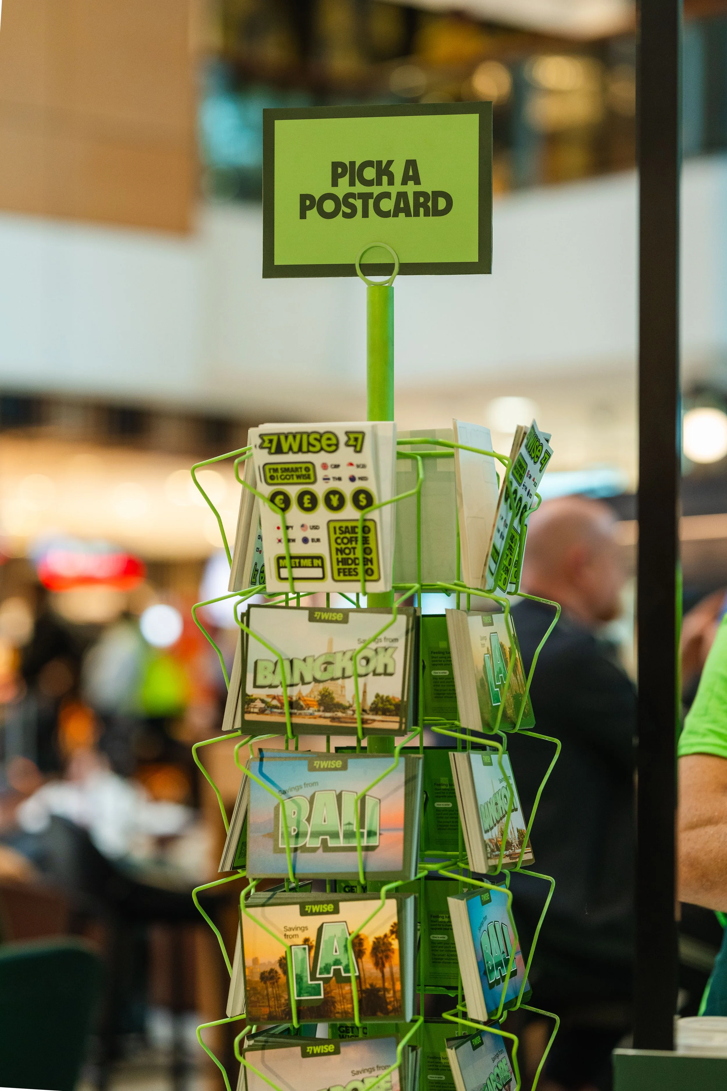 Green postcard display rack with postcards about Bangkok, Bali, and Los Angeles in a public place, with a sign above that says 'Pick a Postcard'.