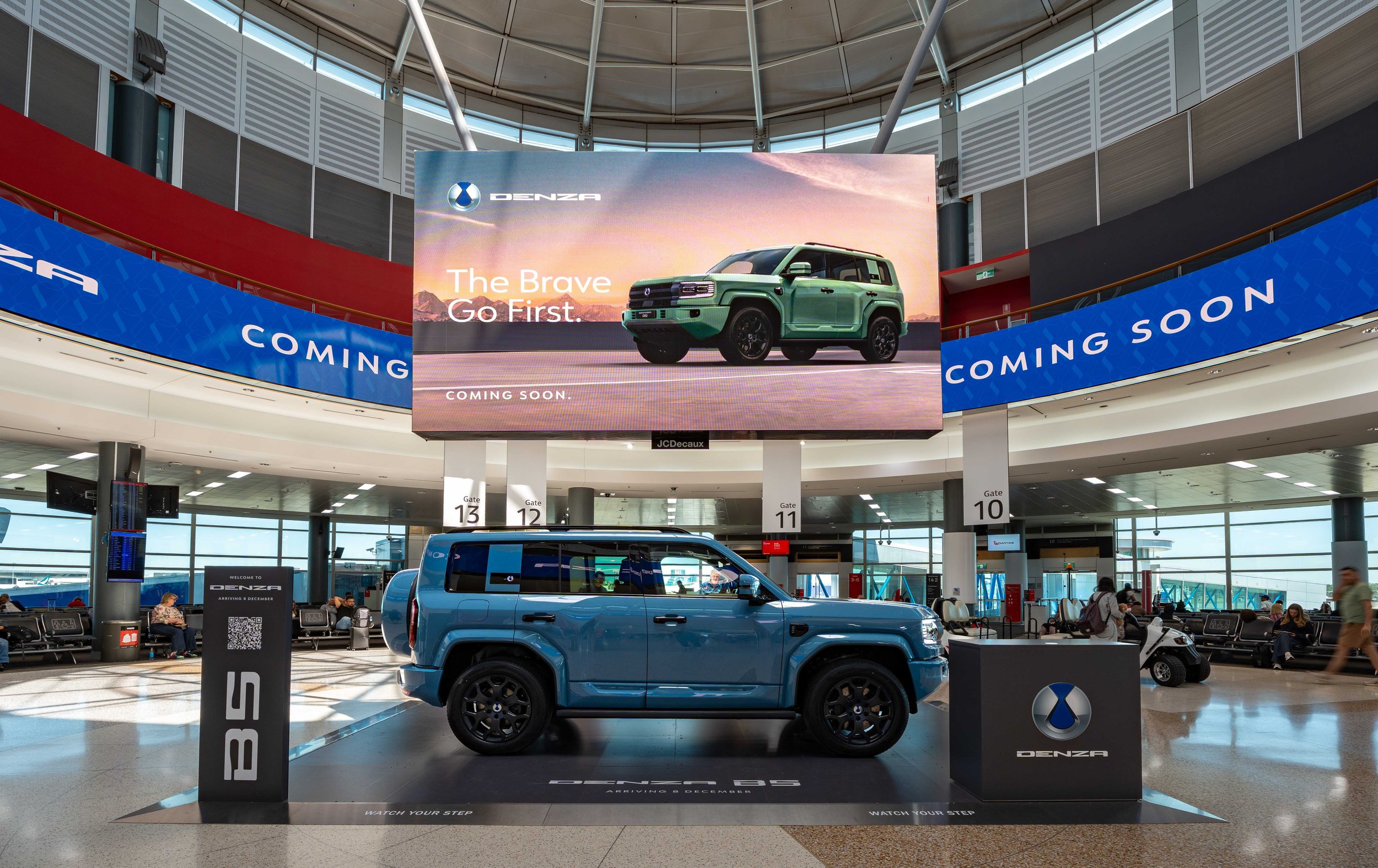 Denza B6 electric vehicle on display at an airport terminal with a large digital billboard showing a different Denza vehicle and the words "The Brave Go First. Coming Soon."