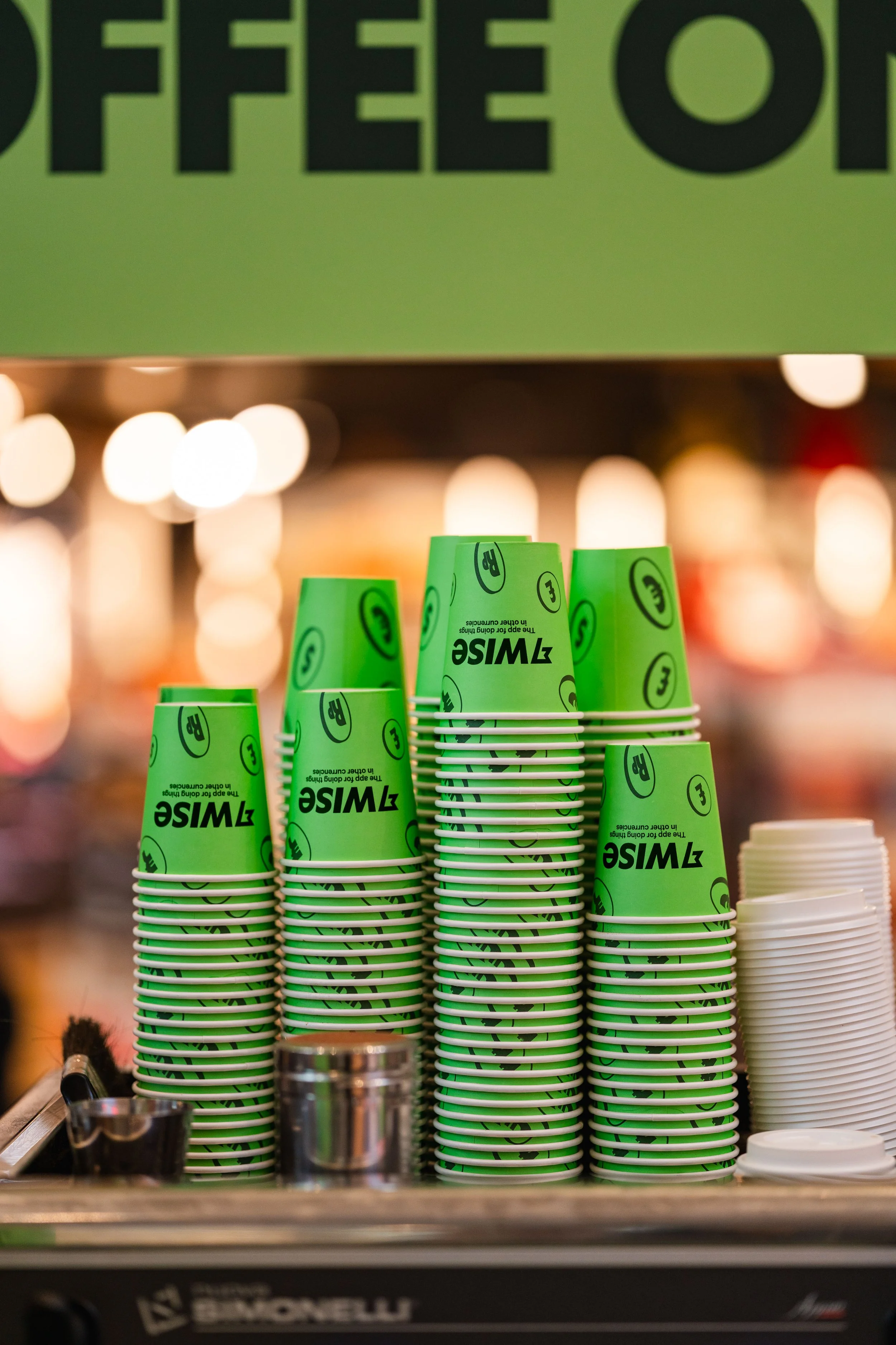 Stacks of green paper coffee cups with the WisE logo on a coffee shop counter, with a blurry background and a green sign above.