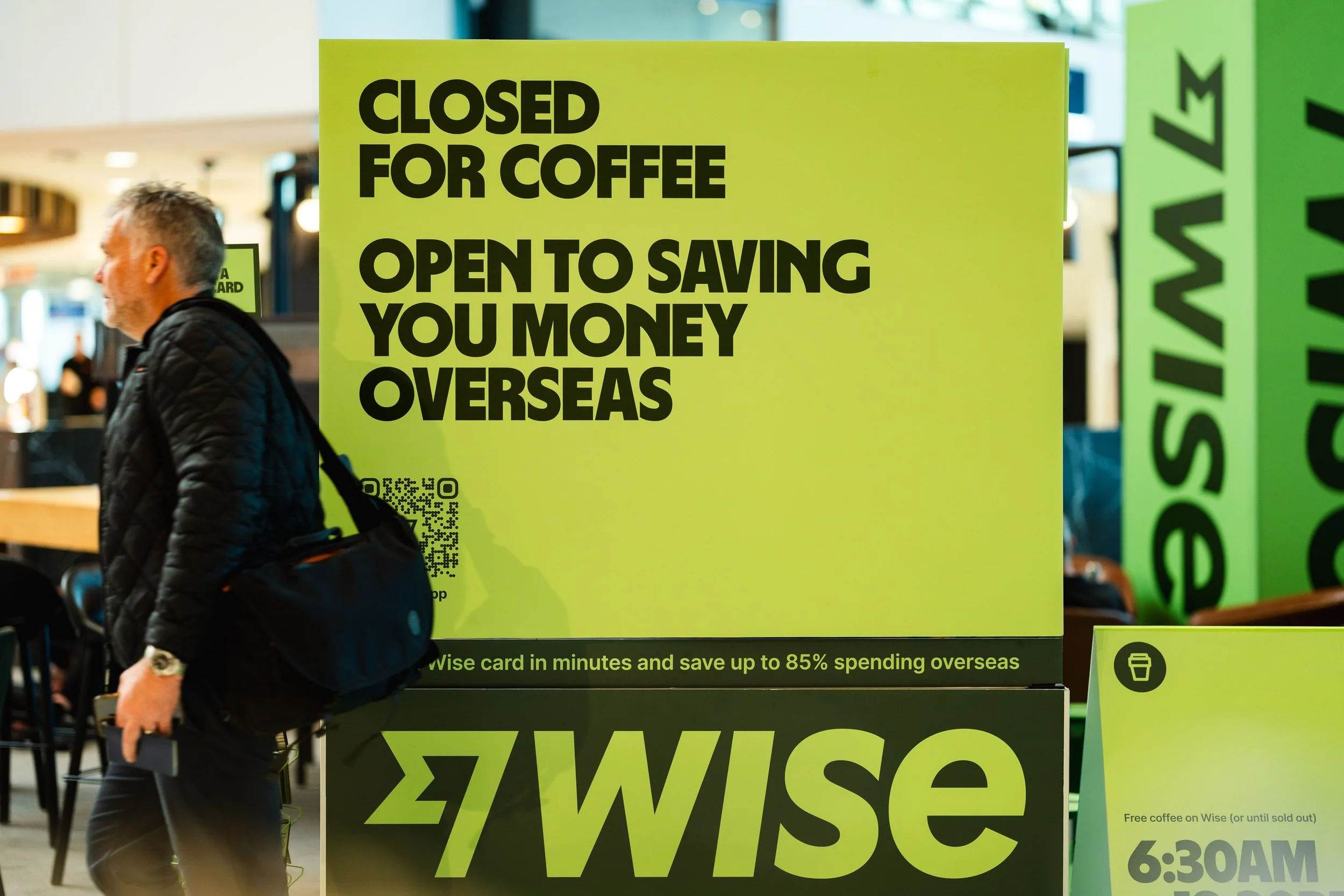 Large yellow sign in an airport or shopping mall that reads "CLOSED FOR COFFEE OPEN TO SAVING YOU MONEY OVERSEAS" with a QR code on the lower left and the "Wise" logo at the bottom. A man with gray hair in a black quilted jacket is walking past the s