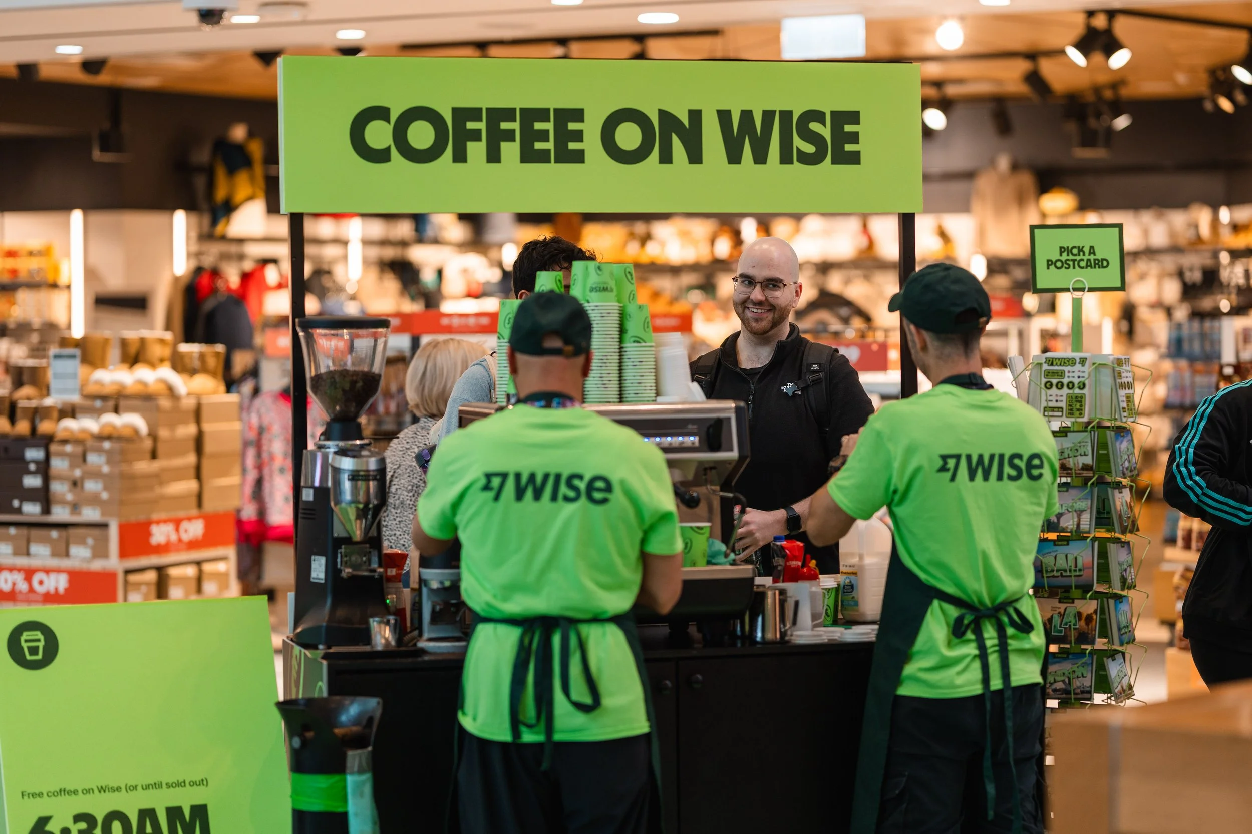 A coffee stand in a store with a large green sign that reads "Coffee on Wise". Two employees wearing bright green shirts with "Wise" on the back prepare coffee, while a smiling customer interacts with them. The background shows shelves with products 