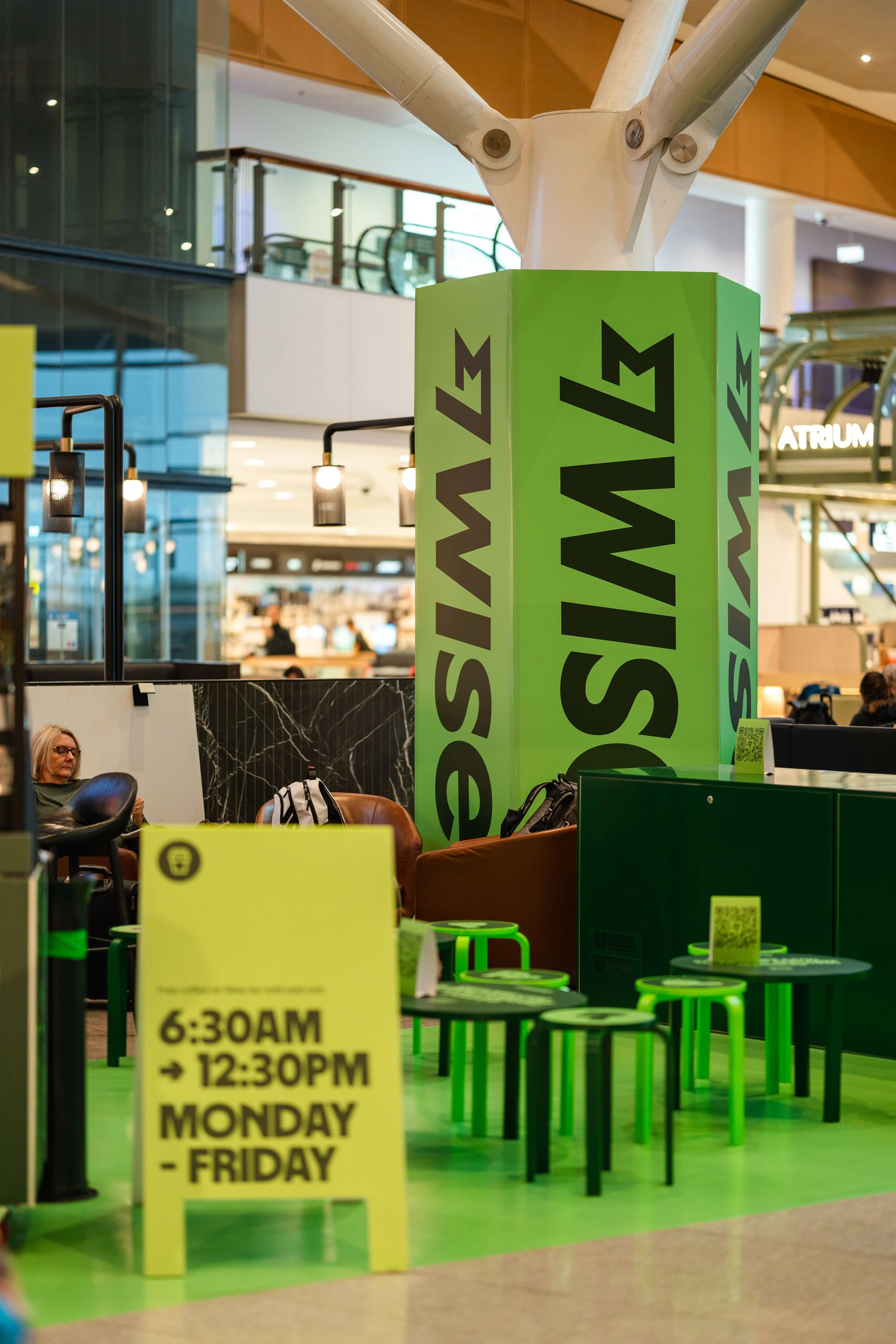 Inside a modern airport, there is a green pillar with large black text that reads 'SWIS' and part of a logo visible. In front, there is a yellow sign indicating the operating hours from 6:30 AM to 12:30 PM, Monday to Friday. The seating area has smal