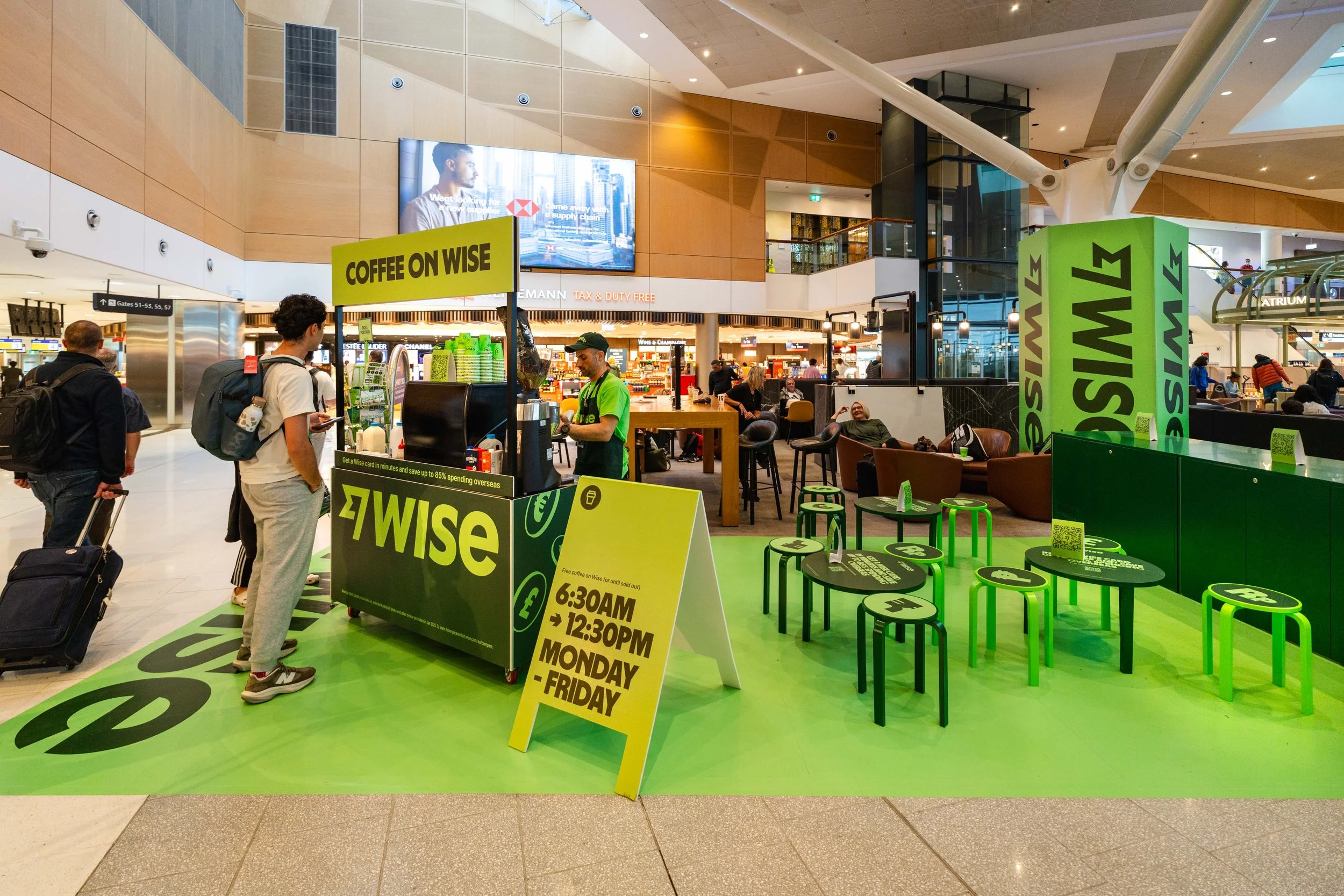 A coffee stand at an airport with a bright green and black color scheme. The sign displays 'Coffee on Wise' and the operating hours from 6:30 AM to 12:30 PM, Monday to Friday. Customers are lining up to order as a barista prepares drinks. The seating