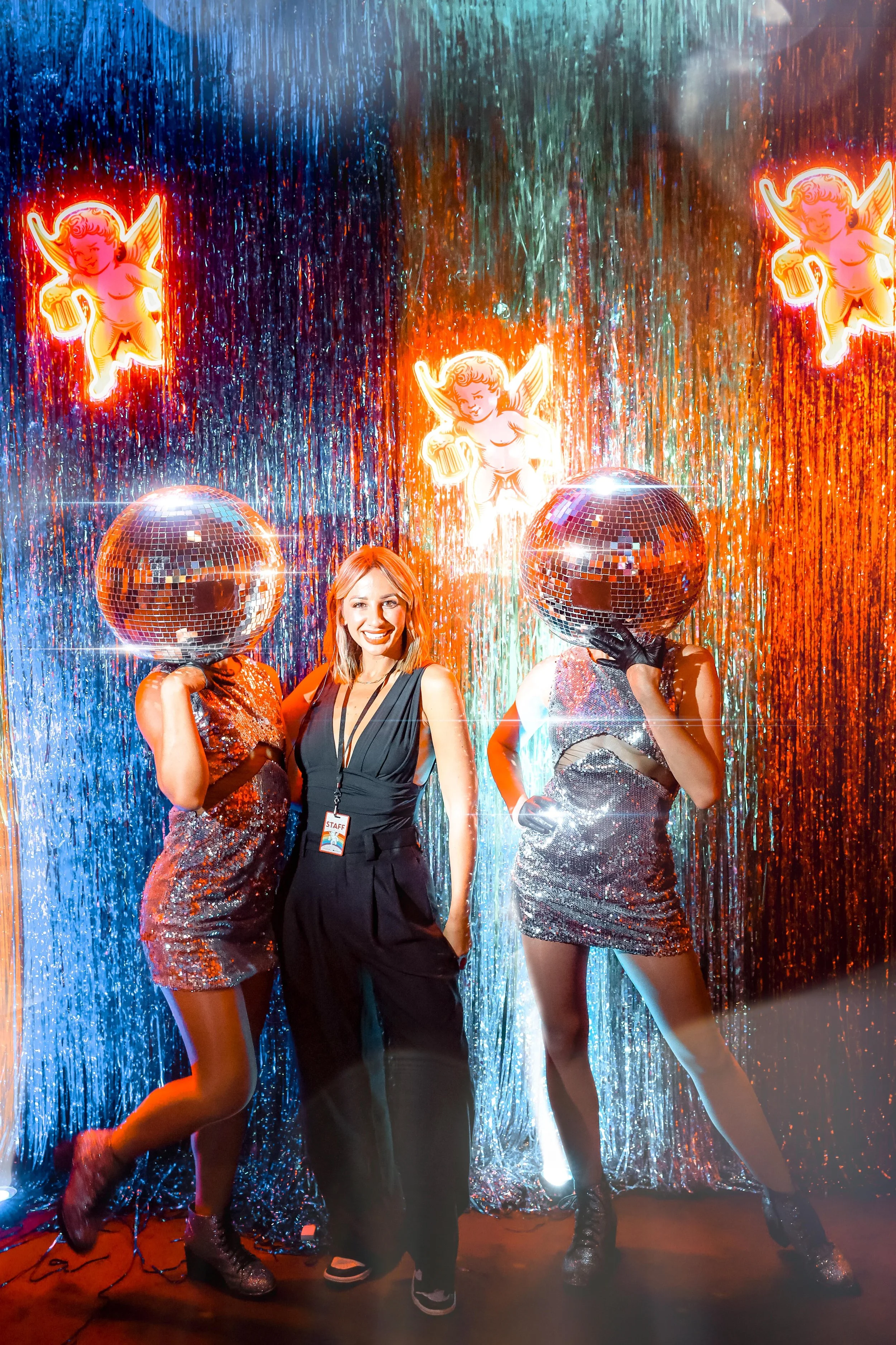 Two women with disco ball helmets and sequined dresses pose with a smiling woman standing between them at a retro-themed party with neon cherub decorations and a glittery backdrop.