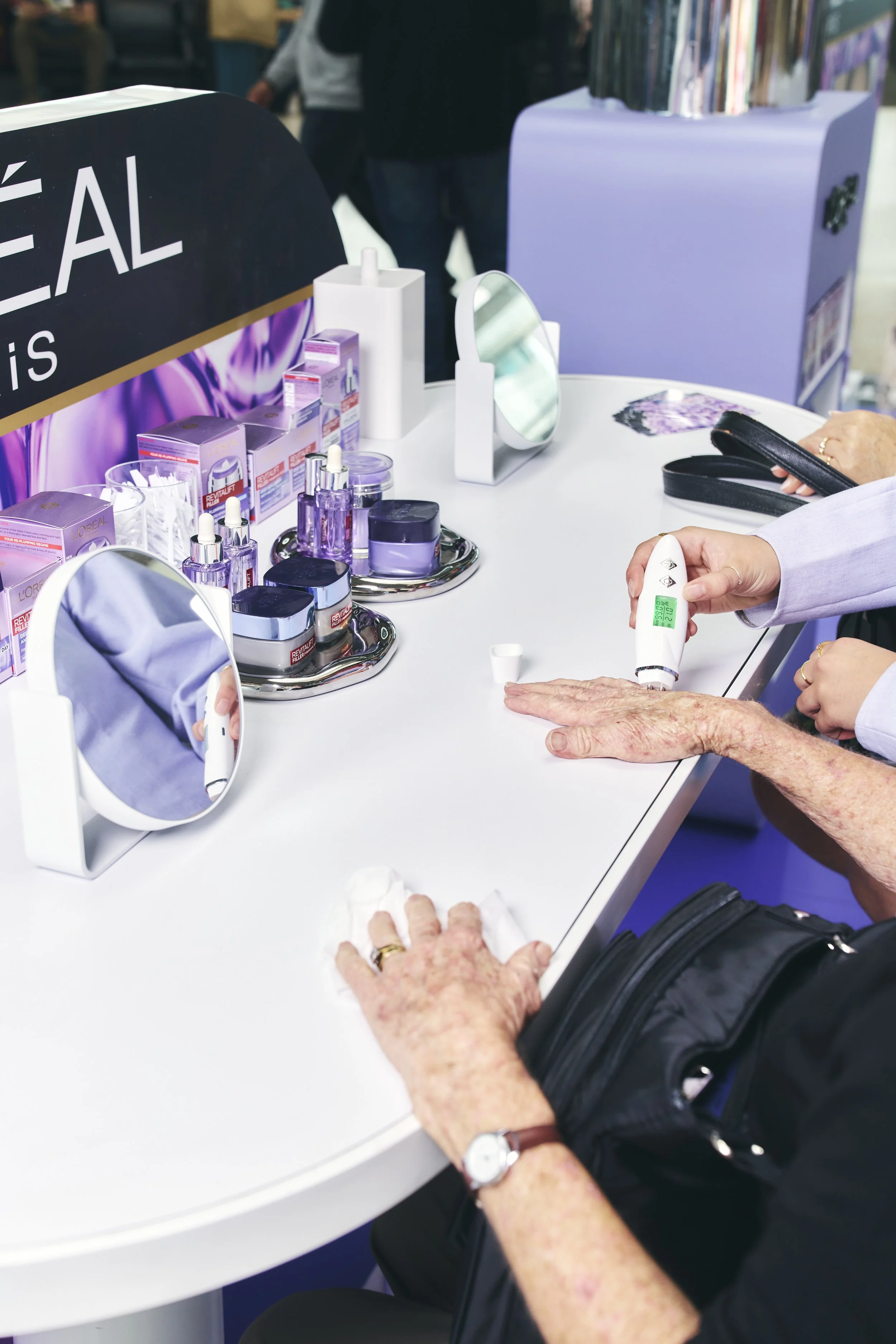 A person receiving a digital skin assessment using a handheld device at a beauty or skincare booth with various skincare products on display.
