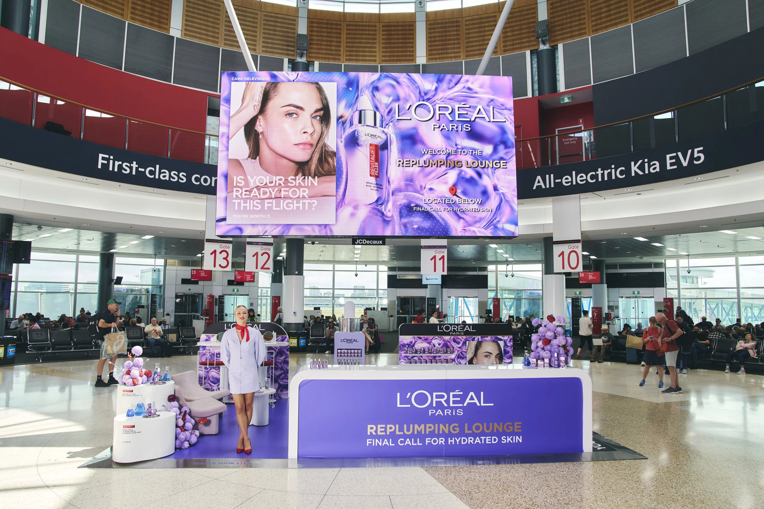 L'Oreal Paris promotional booth at an airport, with a large digital screen advertising skincare products, and a woman standing in front of the booth dressed in a white outfit with red shoes. The booth features lavender and purple decorations, and the