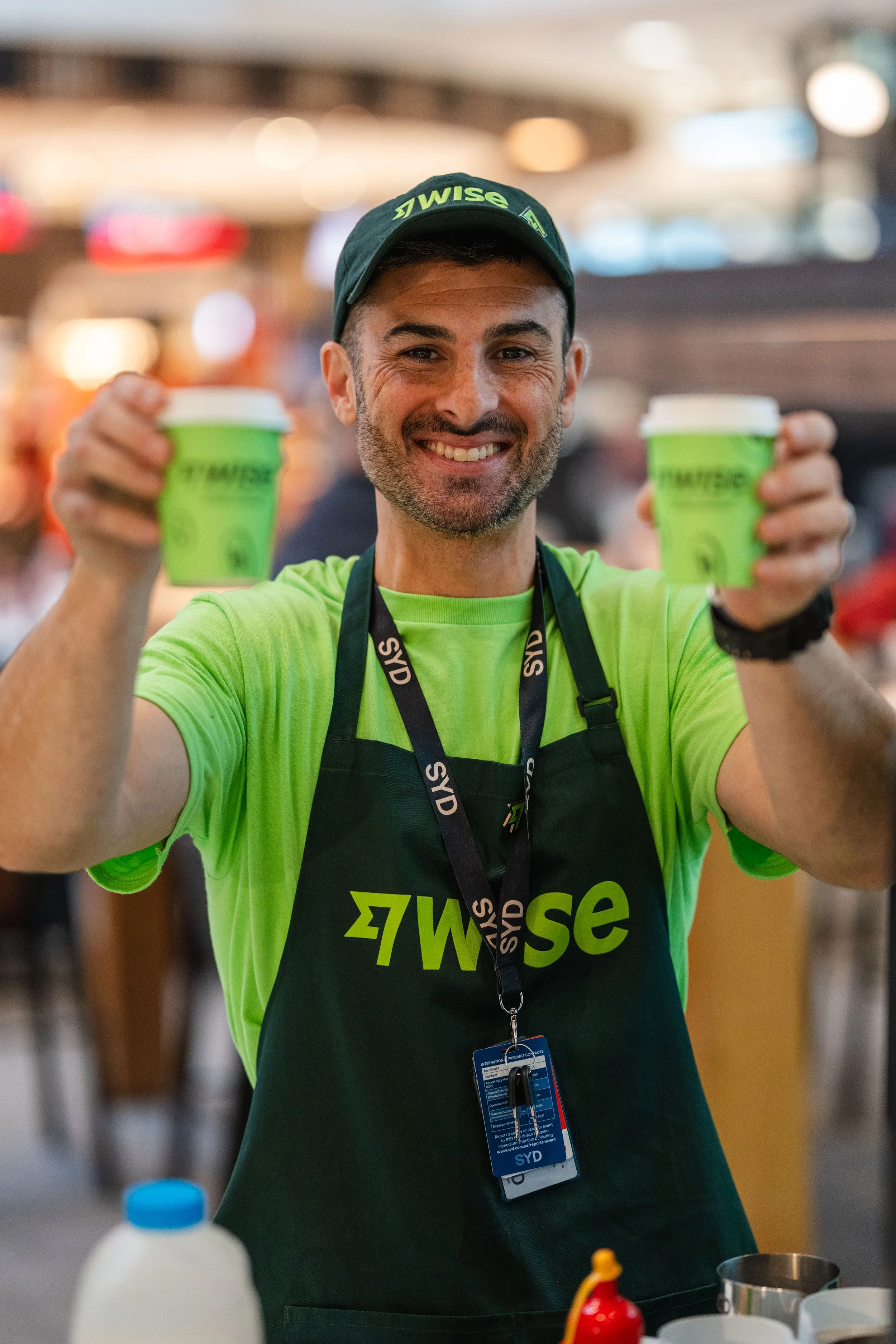 A smiling man wearing a green T-shirt, black apron with 'WISE' written on it, black lanyard with 'SYD' tags, and a cap, holding two cups of coffee in a busy indoor setting.