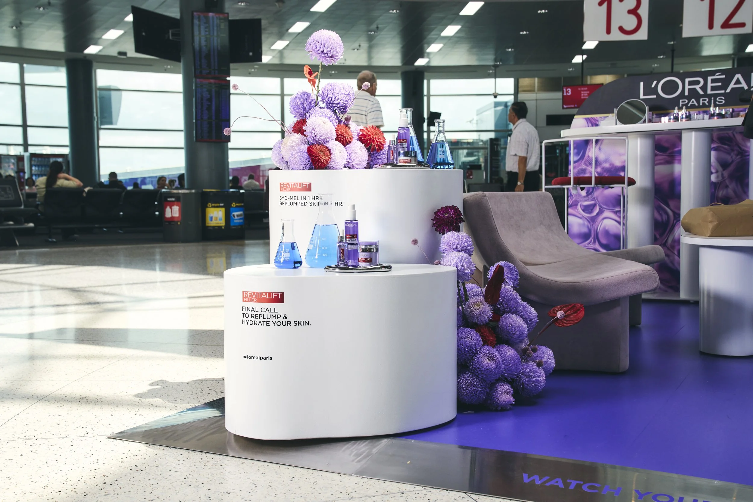 Display at an airport with skincare products, flowers, and a lounge chair, with people and windows in the background.