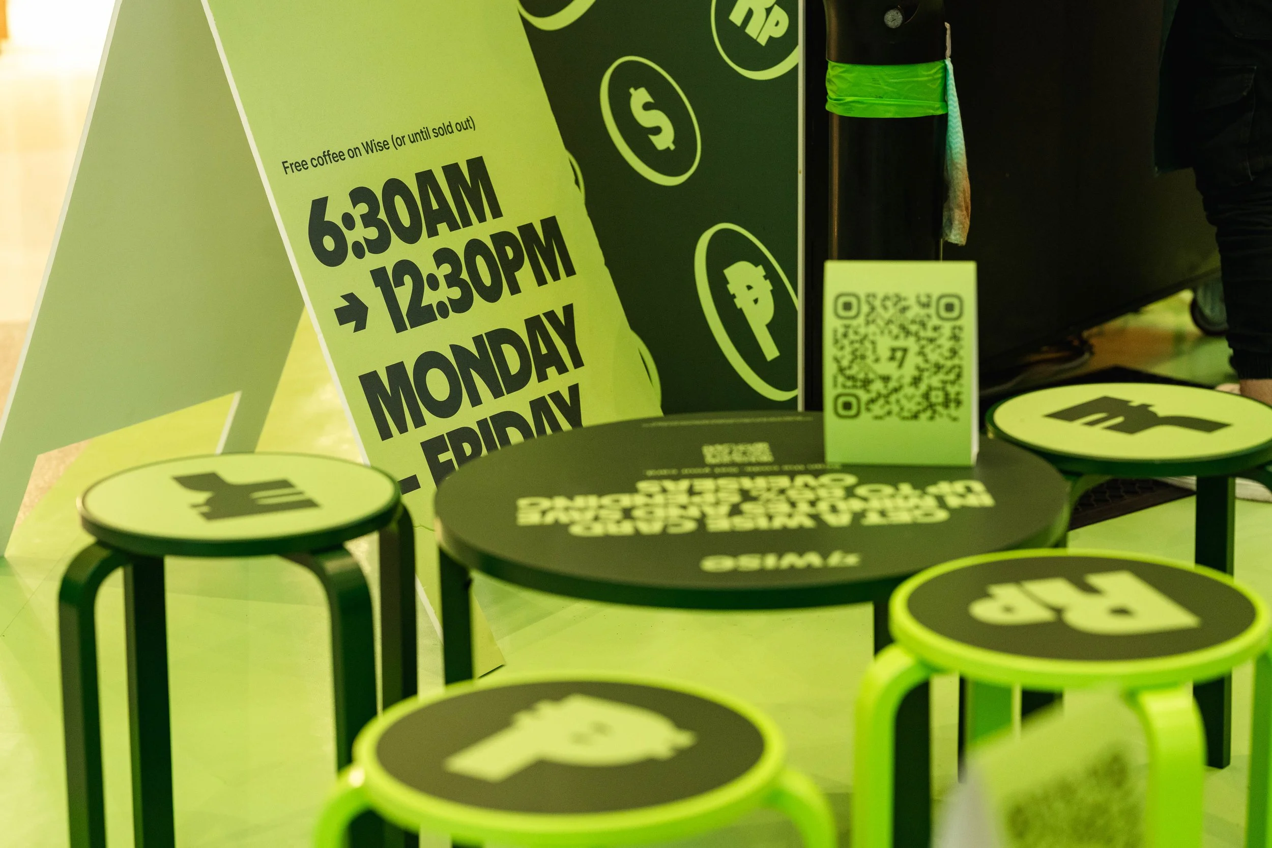 Green and black tables with tabletops displaying a QR code and text, placed in front of a large green sign with business hours from 6:30 AM to 12:30 PM, Monday through Friday, and icons of a dollar sign, coffee, and a power plug.