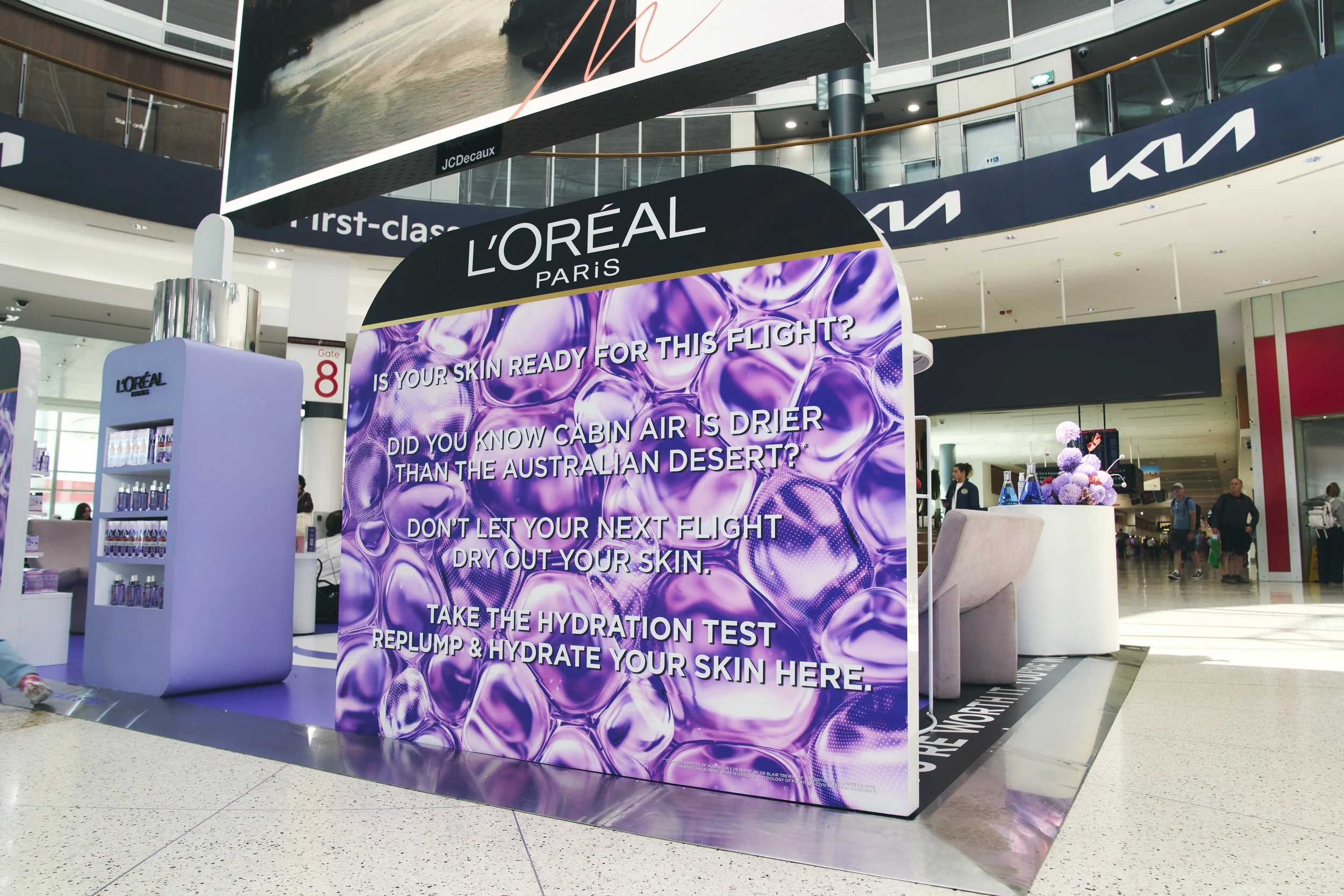 A promotional display for L'Oréal Paris at an airport, advertising a hydration test for skin with bold purple and white text on a decorative purple background.