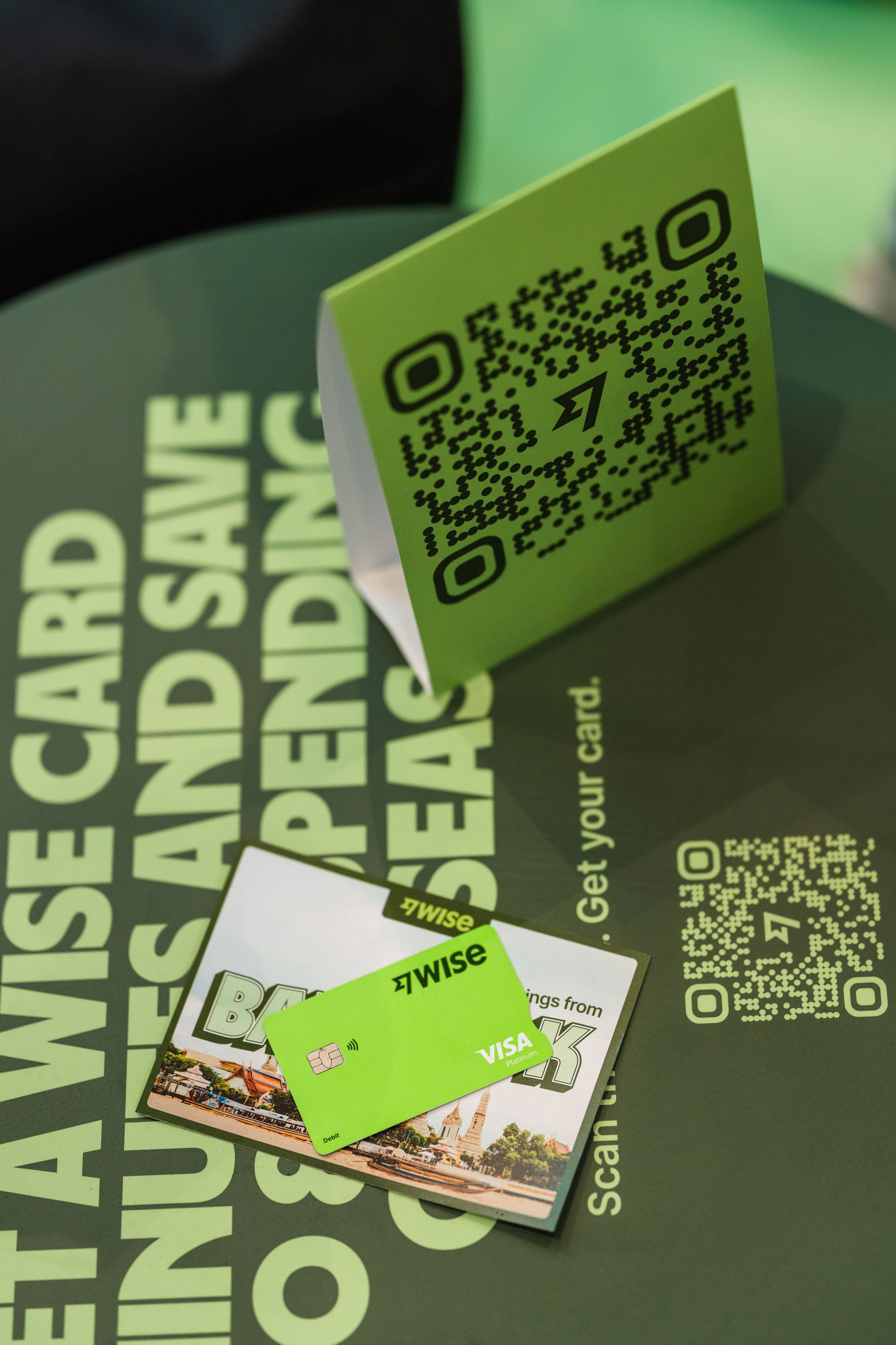 A green Wise Visa debit card placed on a promotional card with Thai landmarks in the background. Behind them, a black sign reads 'WE INSURED CARDS AND SAVINGS ACCOUNT'. There are also QR codes on the sign and a small green folded card.
