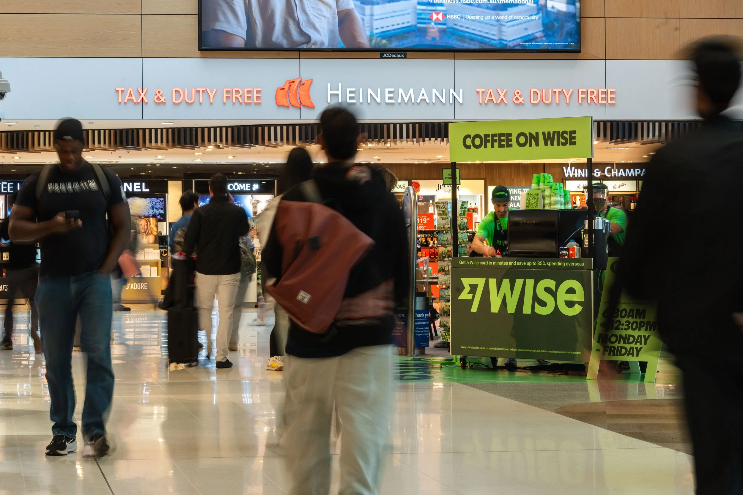 People walking inside an airport terminal near a duty-free shop with 'Heinemann' signage, displaying products and a green Wise card promotion sign.