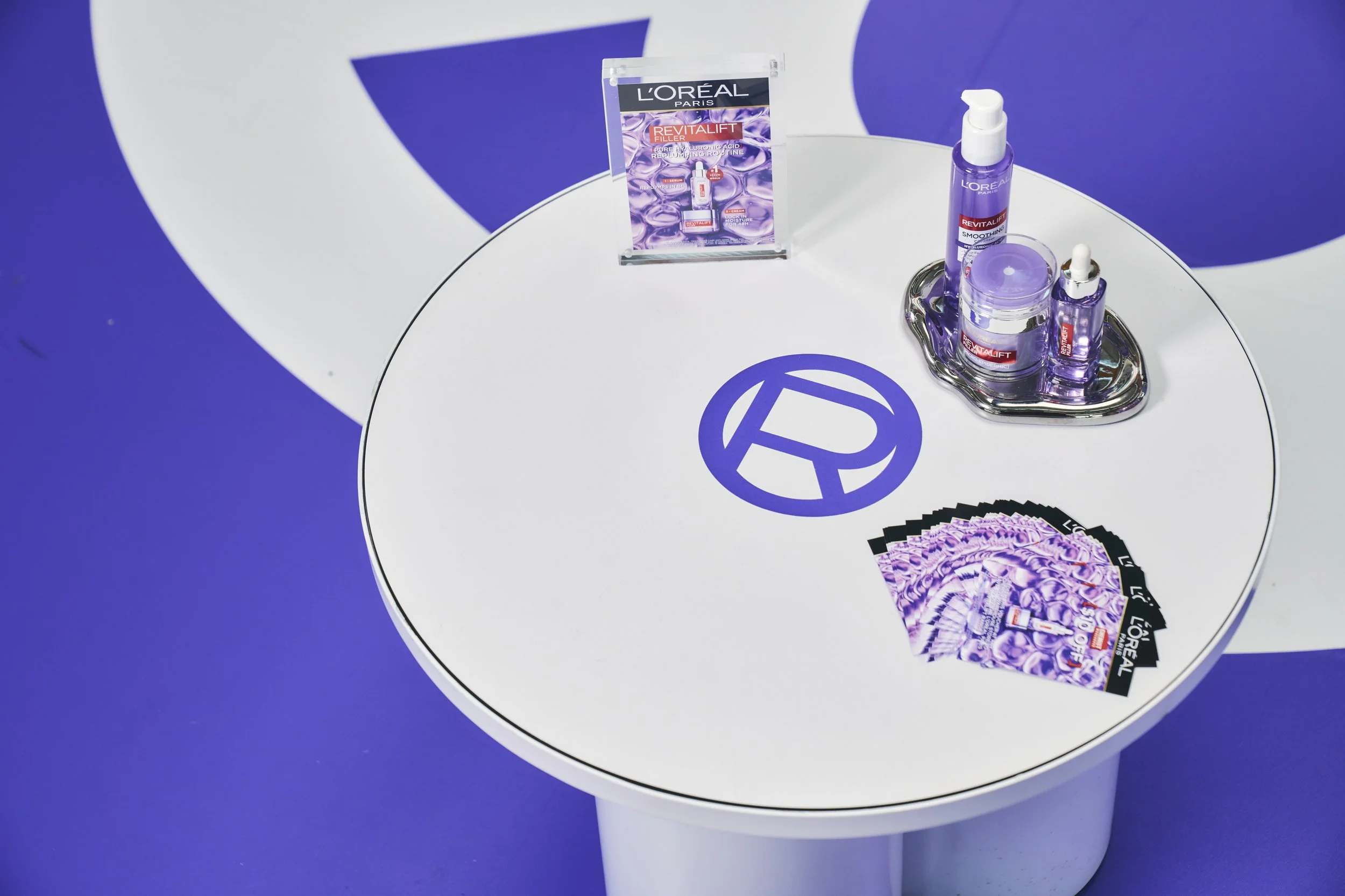 L'Oreal skincare products on a silver tray, promotional flyers, and brochures on a white circular table with purple graphics and branding.
