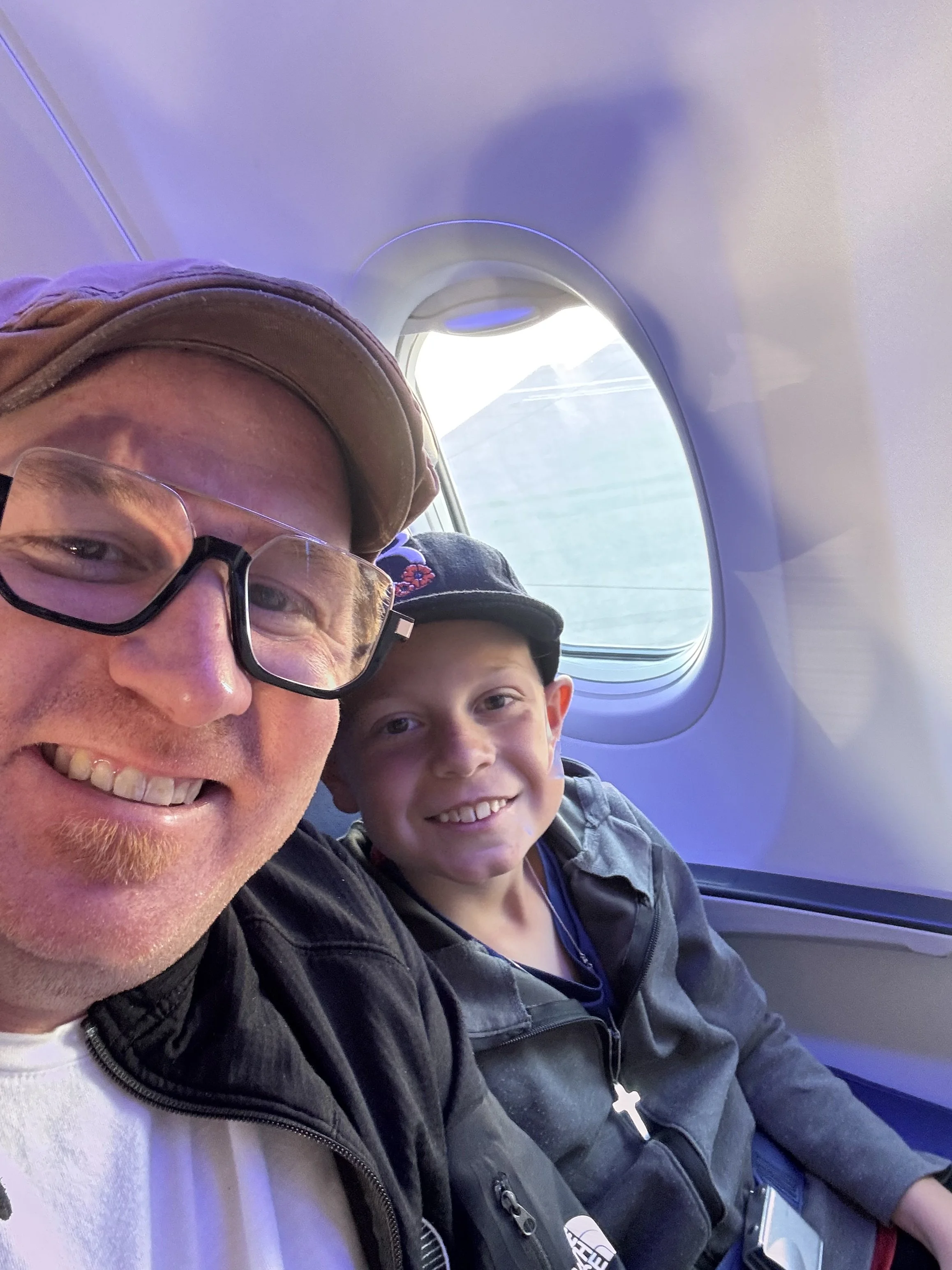 A man and a boy sitting inside an airplane next to a window, smiling at the camera, with the airplane cabin and window visible behind them.
