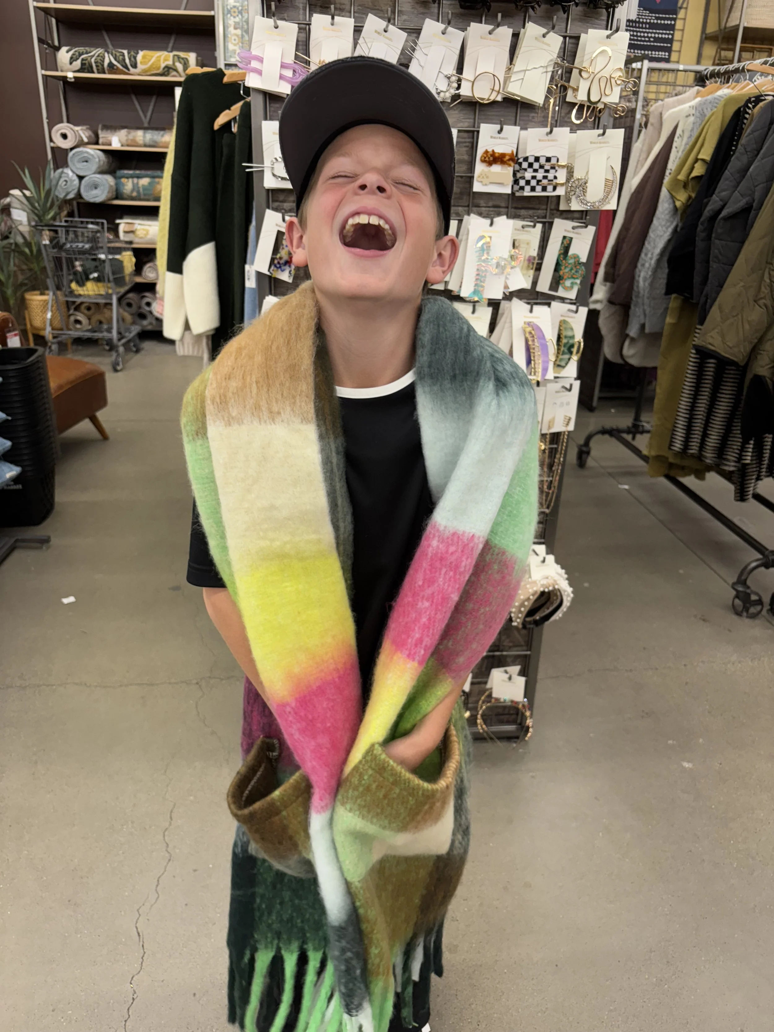 A young boy wearing a black cap and a colorful, striped scarf laughing with his eyes closed inside a store that sells accessories and clothing, with jewelry displays and racks of jackets in the background.