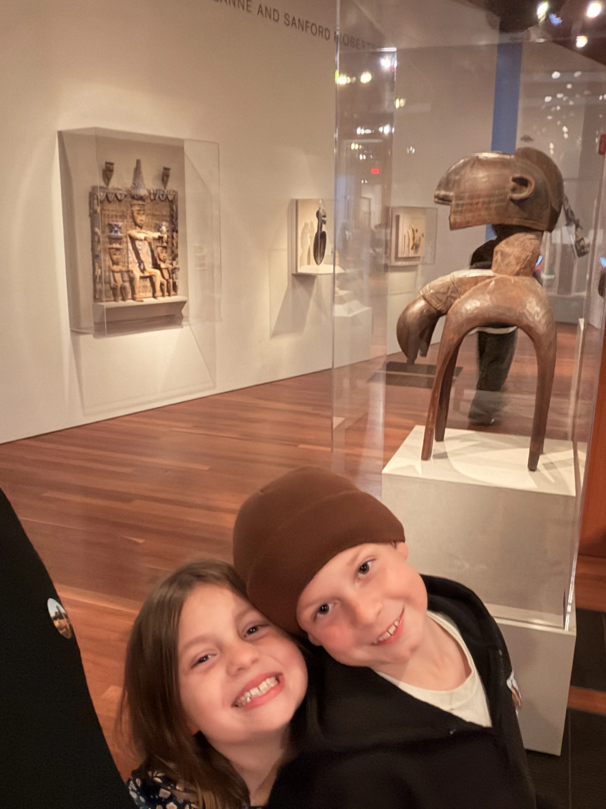 Two young children smiling and posing for a selfie, with museum exhibits visible in the background.