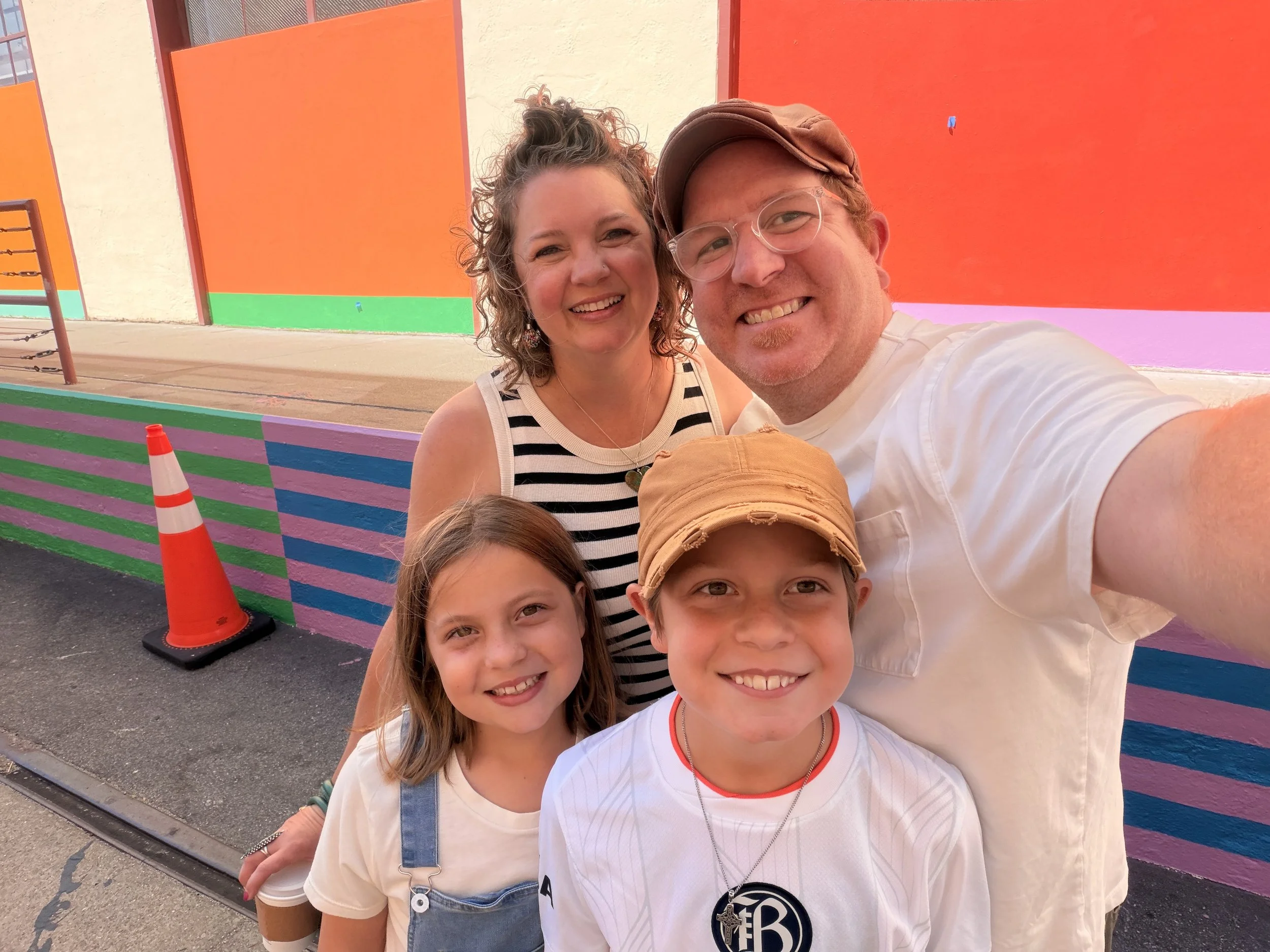 A group of five people taking a selfie in front of a colorful, striped wall with bright orange, green, purple, and pink sections. Two children are in the front, smiling, while two women and a man are behind them, also smiling. The man is wearing glas