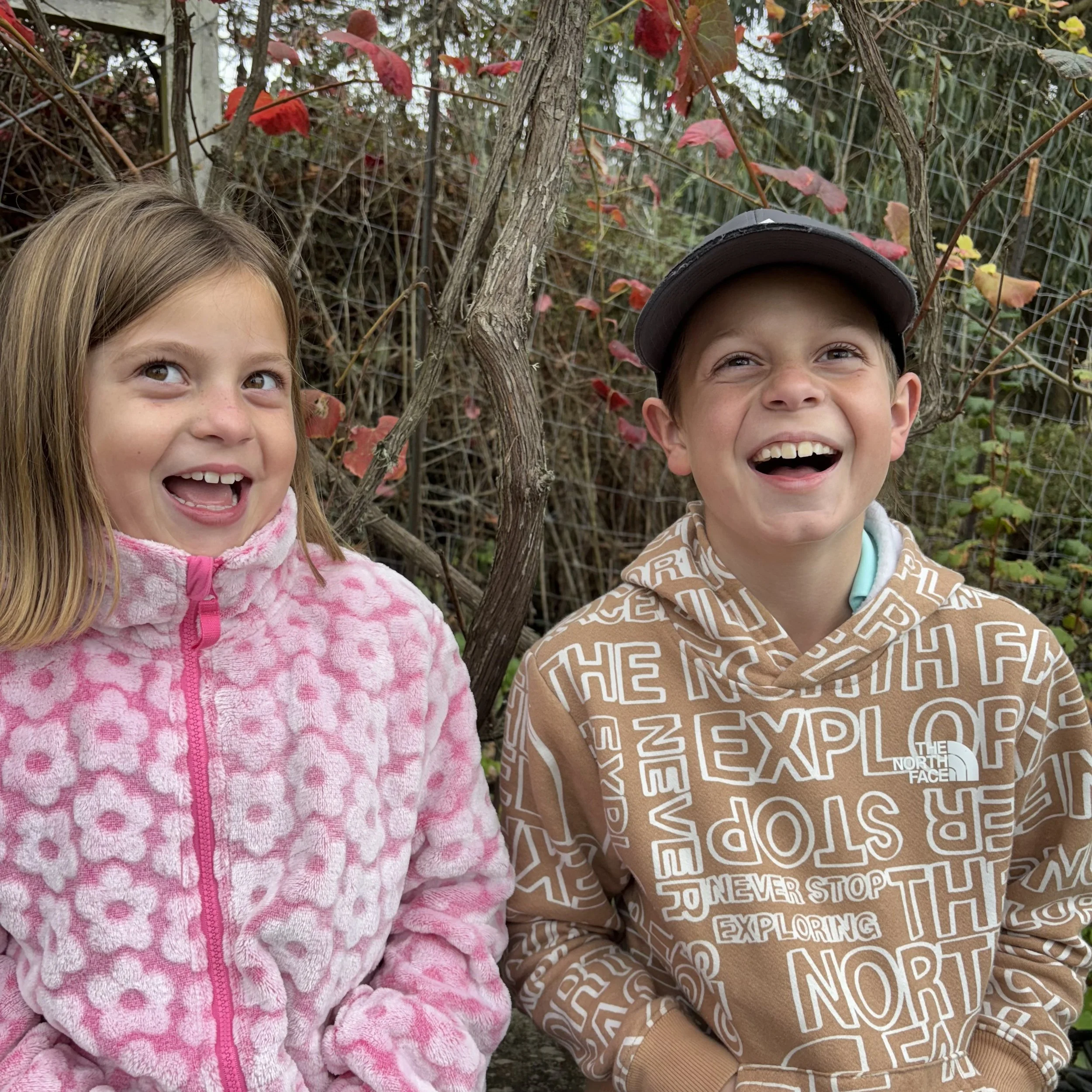 Close-up of two children smiling outdoors with foliage and a wire fence in the background.