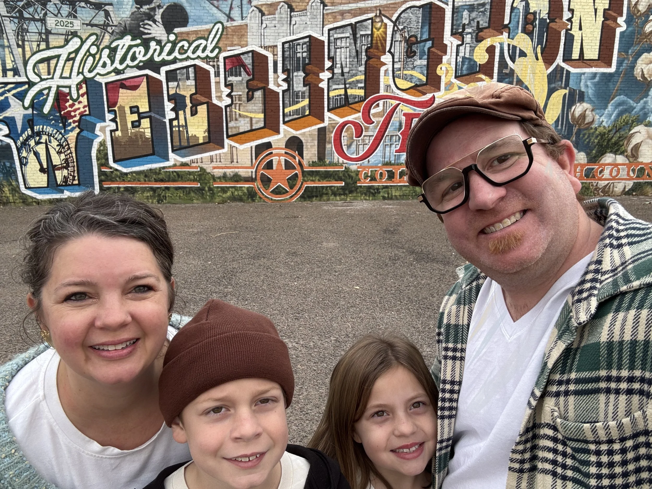 A group of five people, including two women and three children, smiling for a selfie in front of a colorful mural on a brick wall. The mural has the words 'Historical' and 'Fort Worth' and features various images and designs.