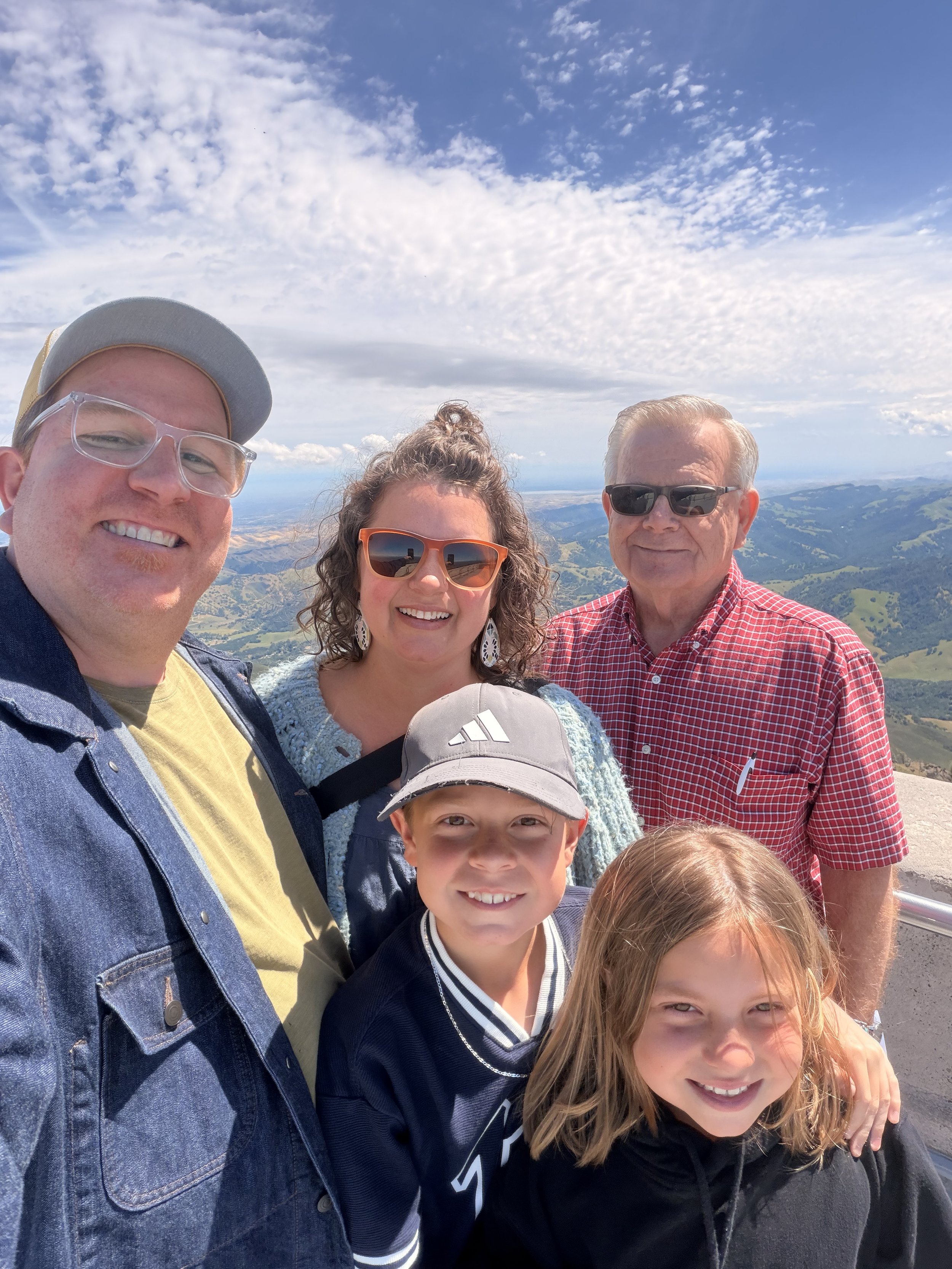 Six people, including children and adults, smiling and taking a group selfie outdoors against a scenic background of rolling hills and a cloudy sky.