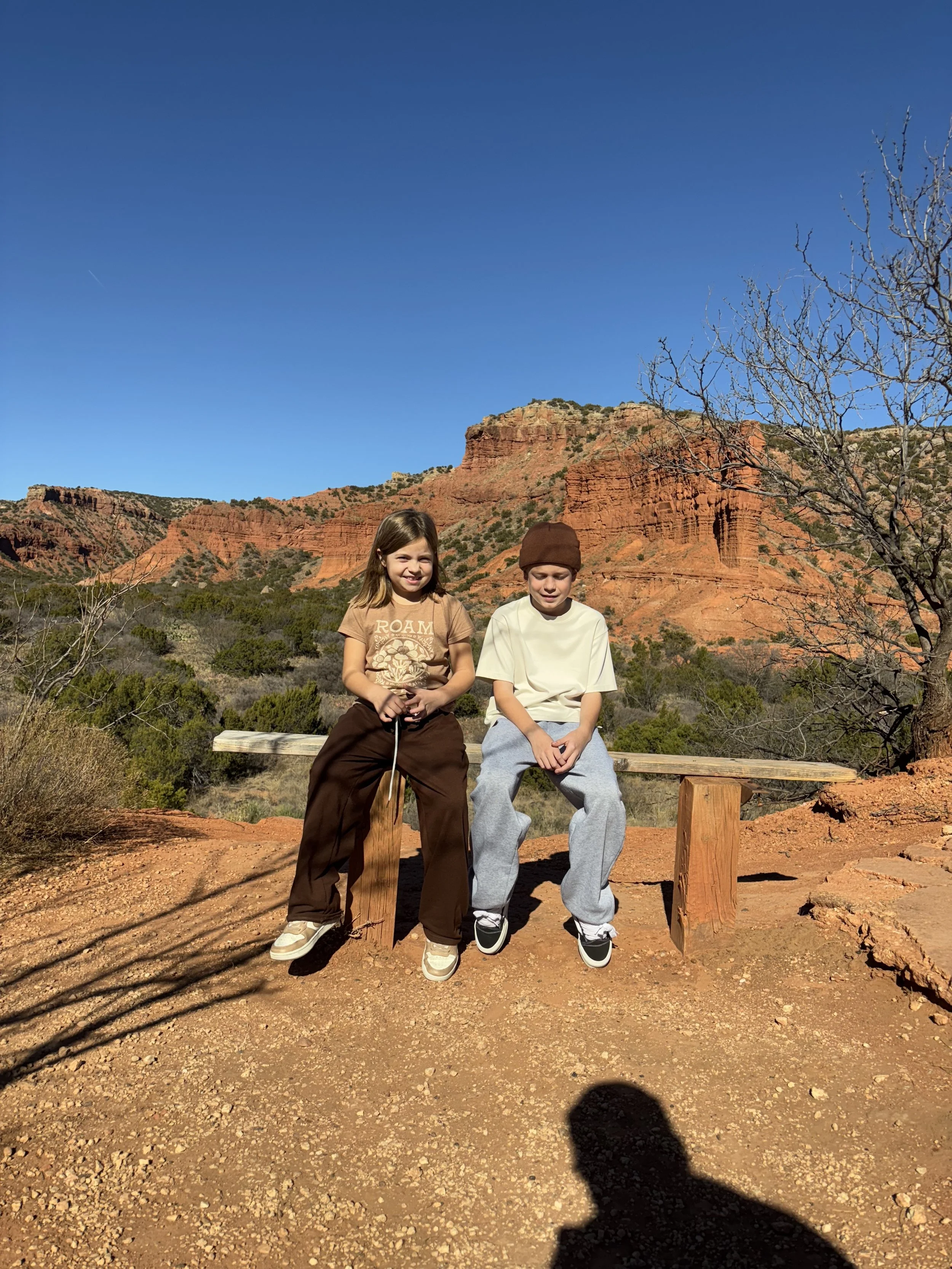 Two children sitting on a wooden bench outdoors in a desert landscape with red rock formations and a clear blue sky.