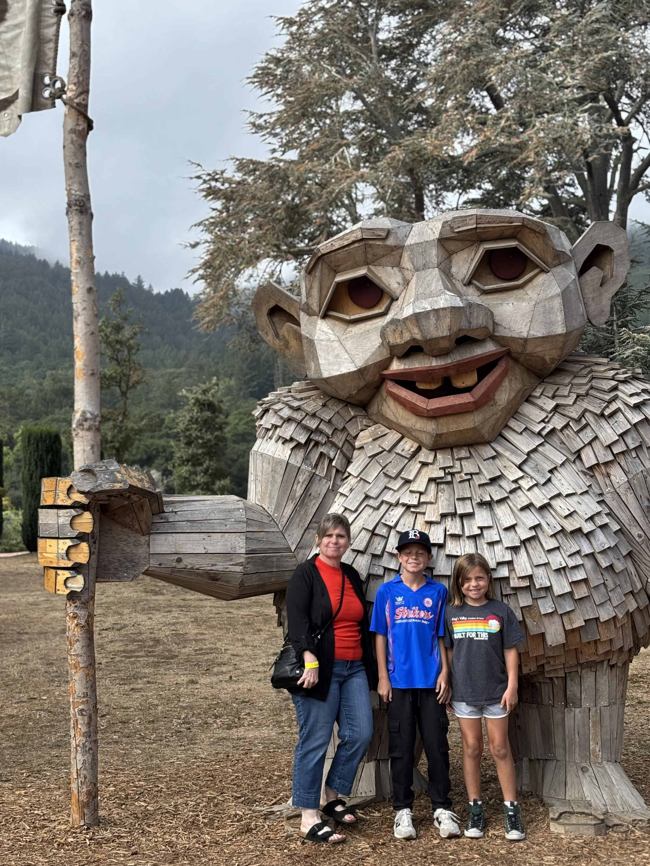 A woman and two children stand in front of a large wooden sculpture of a monkey with a smiling face and big eyes, outdoors in a park with trees and a mountain in the background.