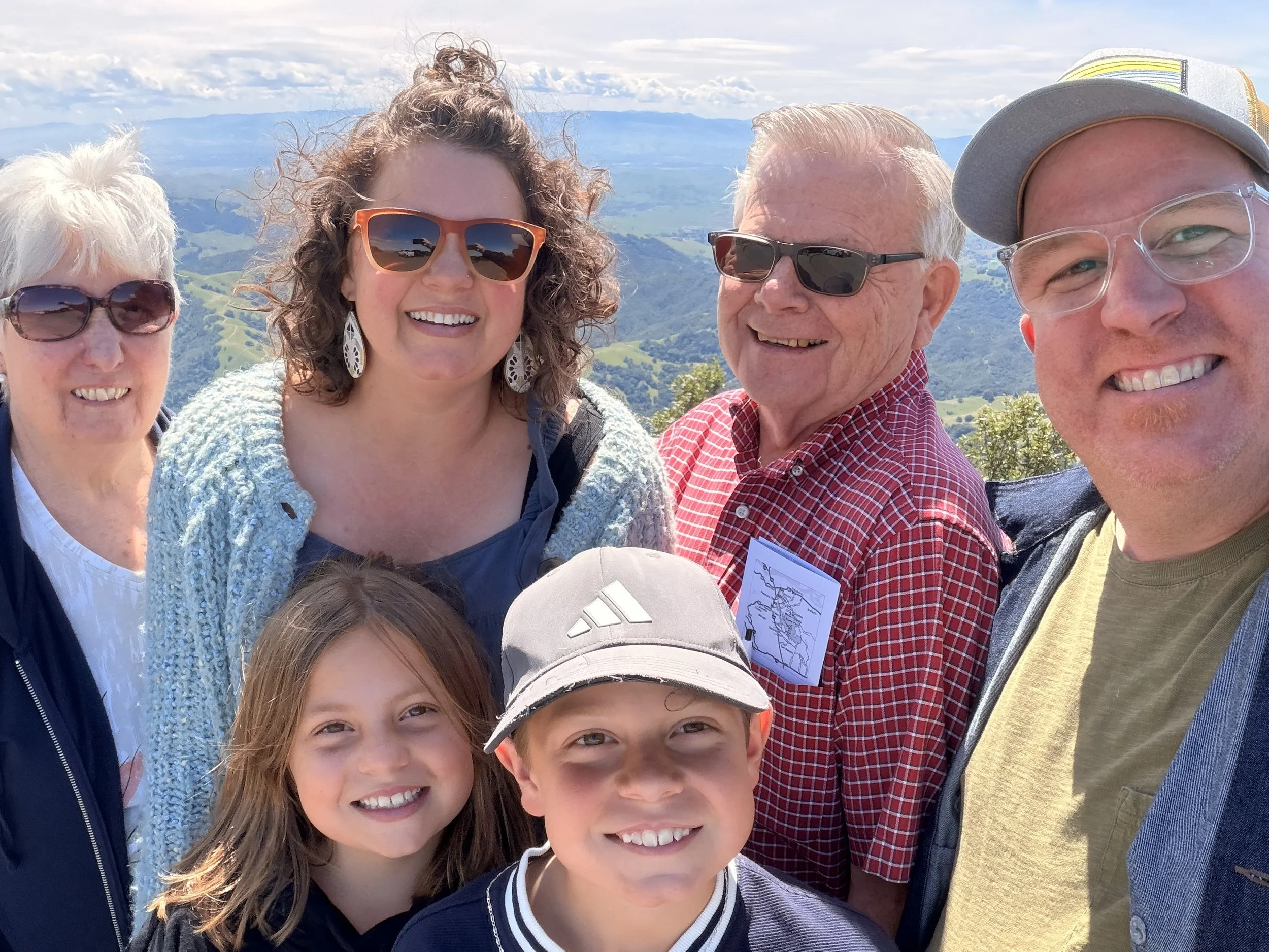 Group of five women and children taking a selfie outdoors with mountain and valley scenery in the background.