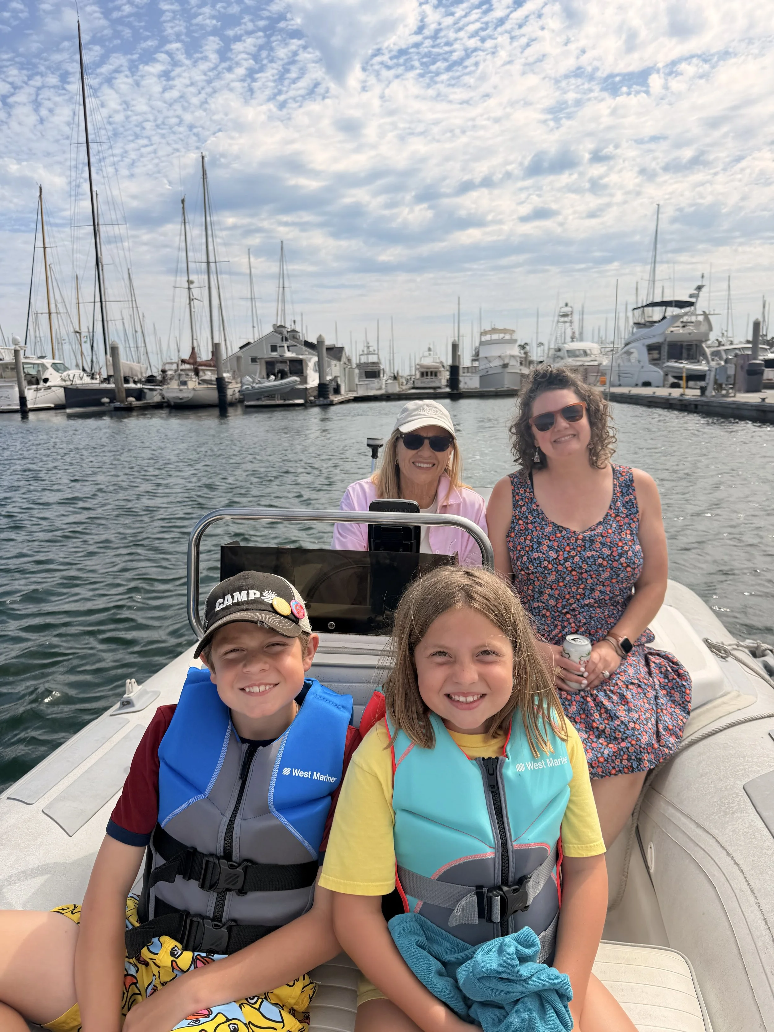 A group of five smiling people on a boat in a marina, with sailboats and yachts docked in the background under a partly cloudy sky.