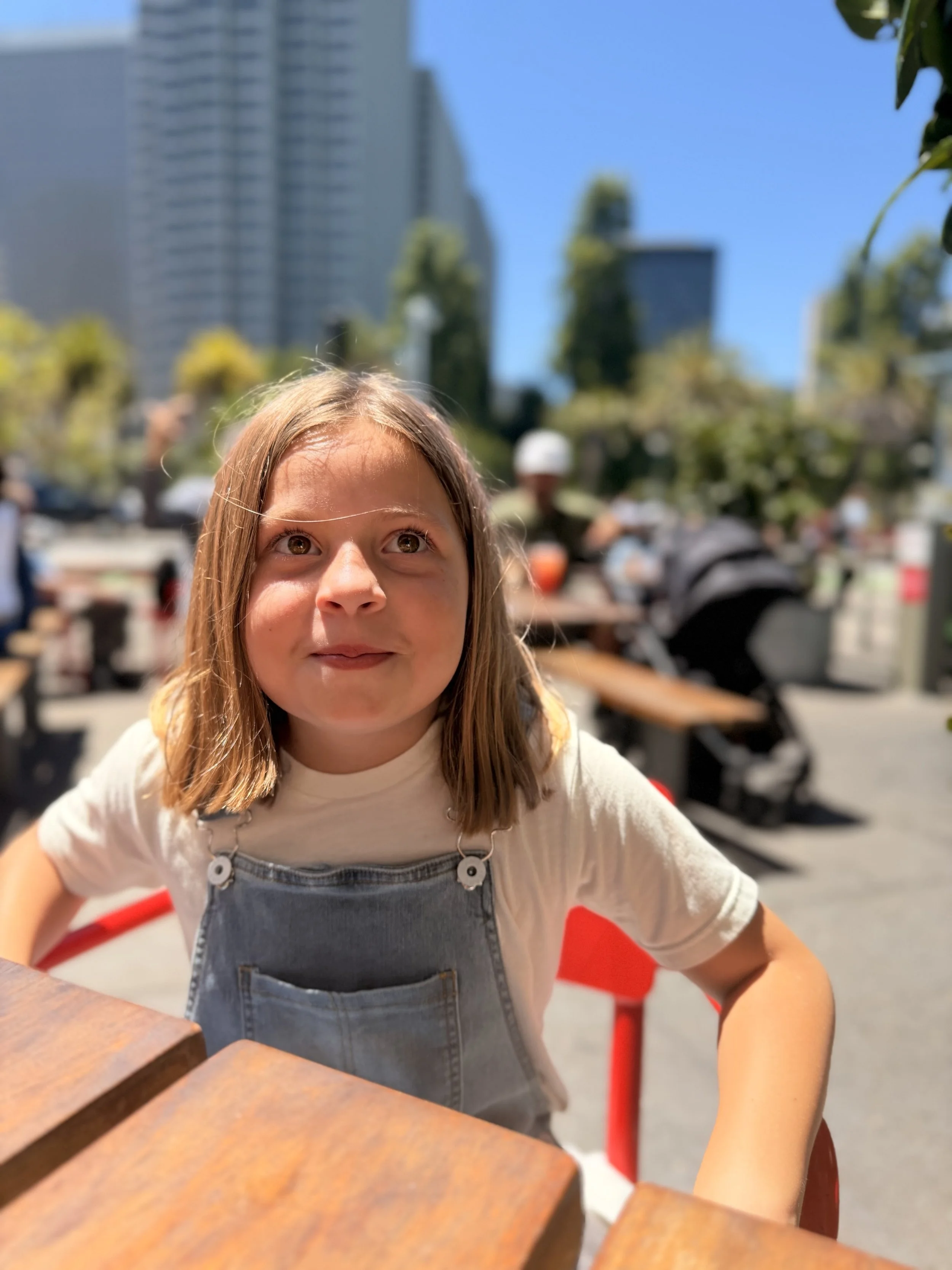 A young girl with light brown hair and brown eyes, wearing a grey denim overall dress over a white t-shirt, faces upward with a slight smile, sitting outdoors on a sunny day with a city skyline and people in the background.