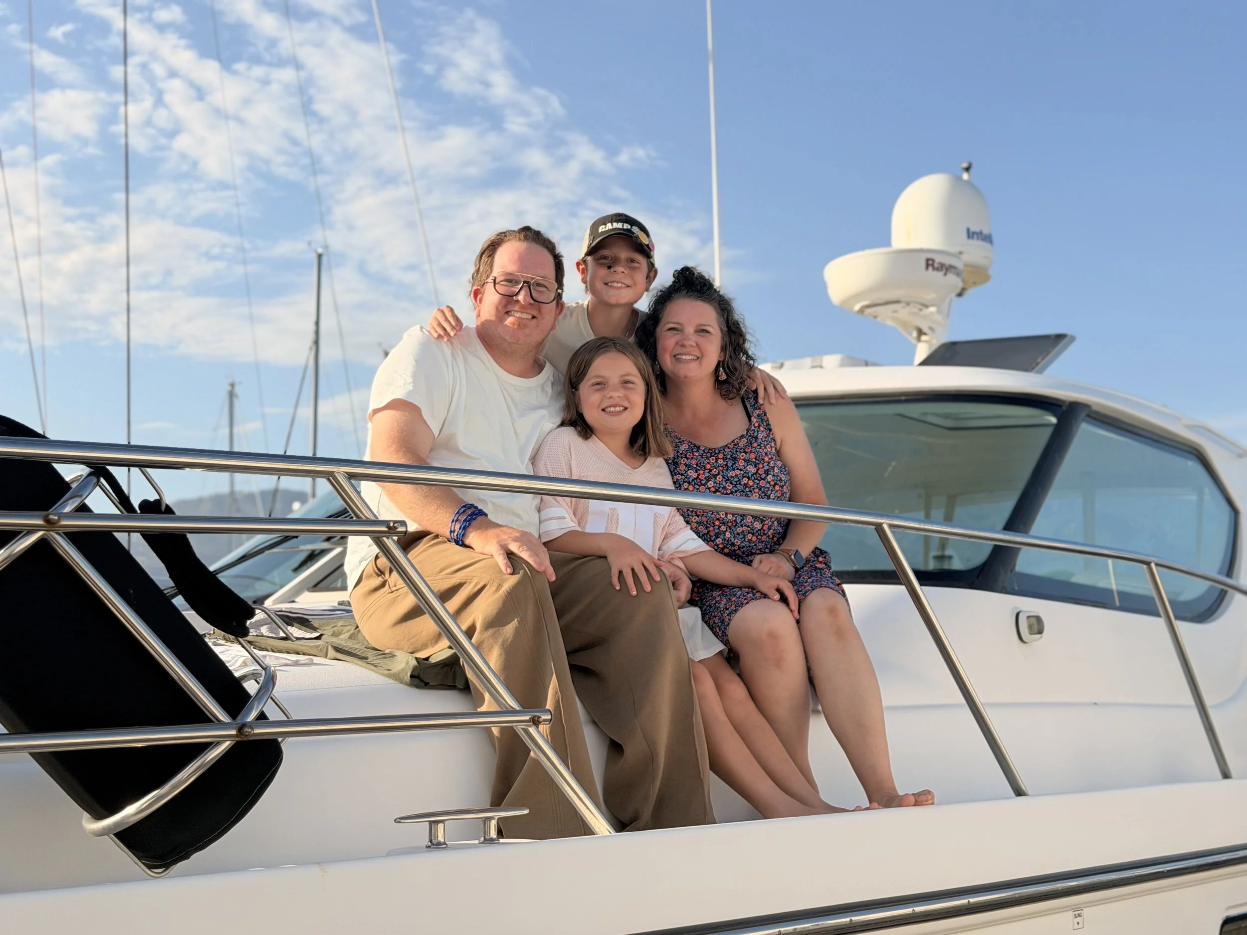 A family of four sitting on the deck of a white boat, smiling outdoors with a clear blue sky and tall antenna equipment in the background.