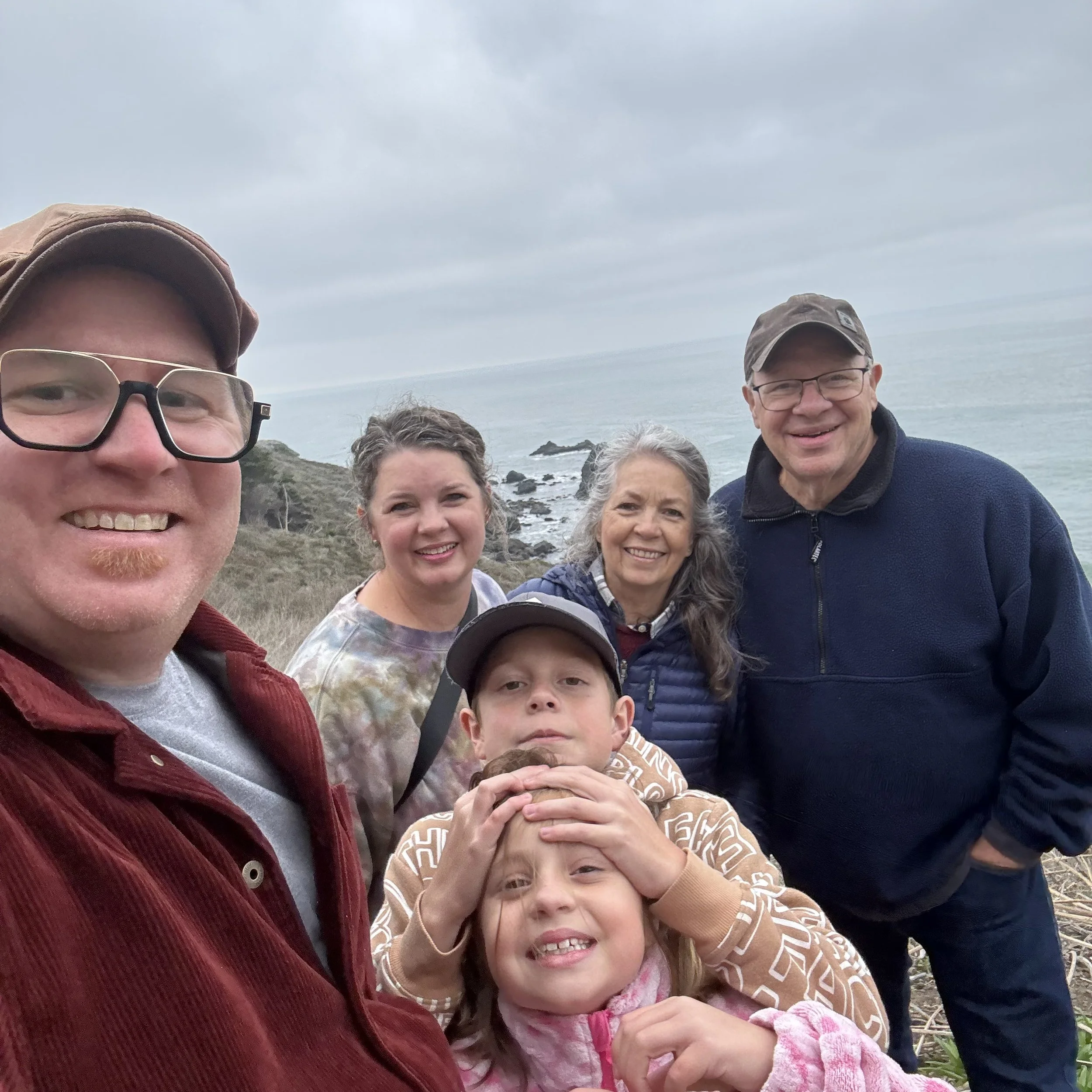 A group of people taking a selfie outdoors by the coast, with rocky shoreline and ocean waves in the background. The group includes adults and children, all smiling.