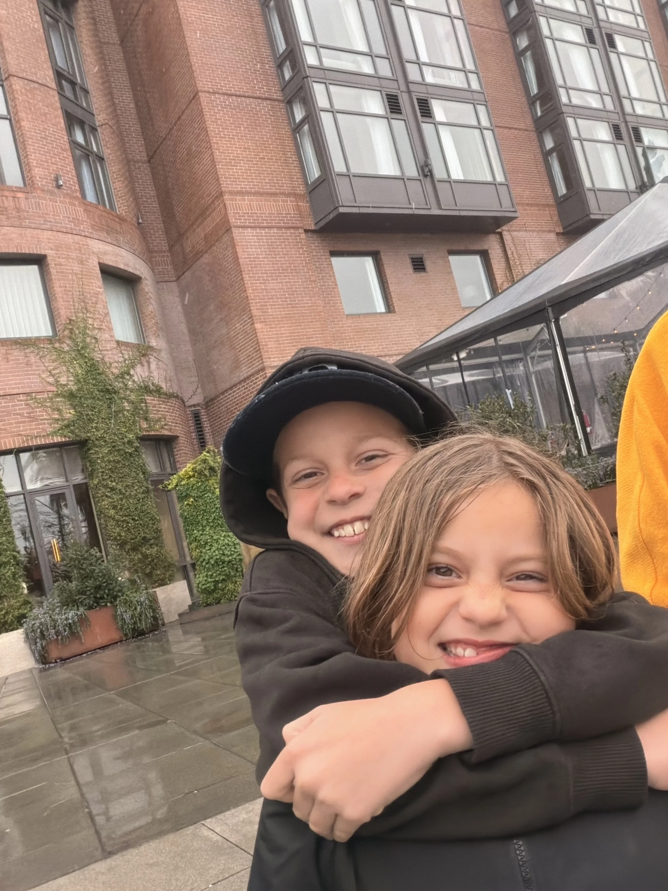 Two children hugging and smiling outdoors in front of a red brick building with large windows and a glass canopy.