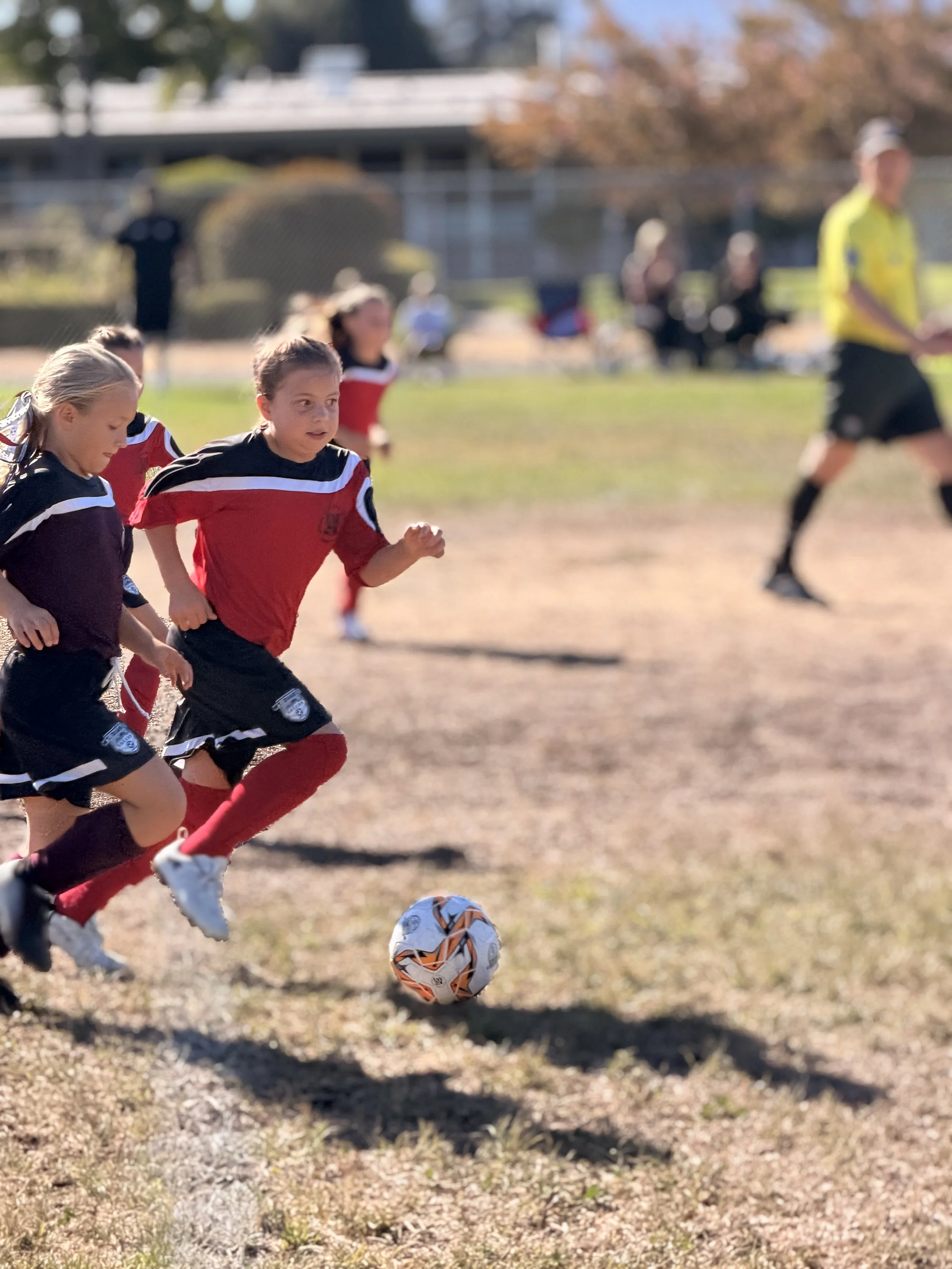 Children playing soccer outdoors on a sunny day, running after a soccer ball on a dirt field with trees and a building in the background.