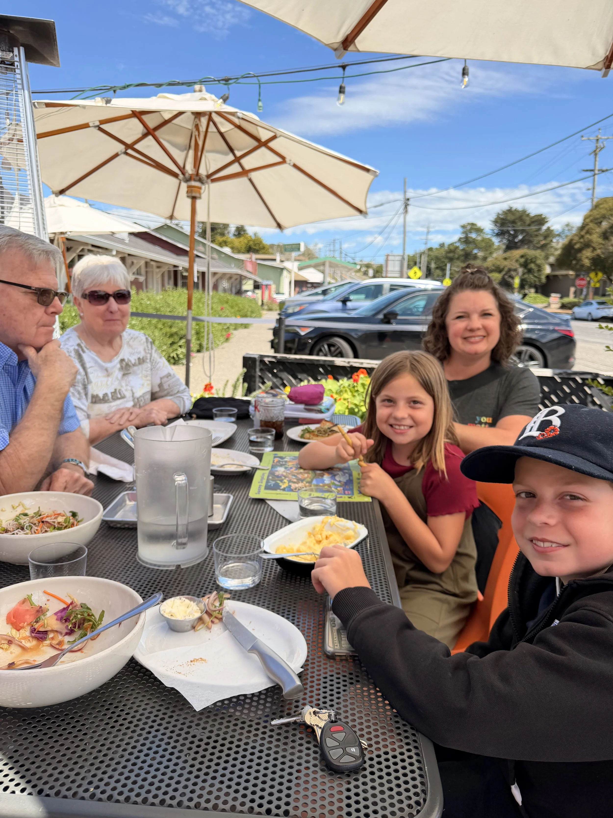 A group of people sitting at an outdoor table with food, including a bowl of salad, mashed potatoes, and drinks. There are umbrellas, string lights, and parked cars in the background, with a clear blue sky.