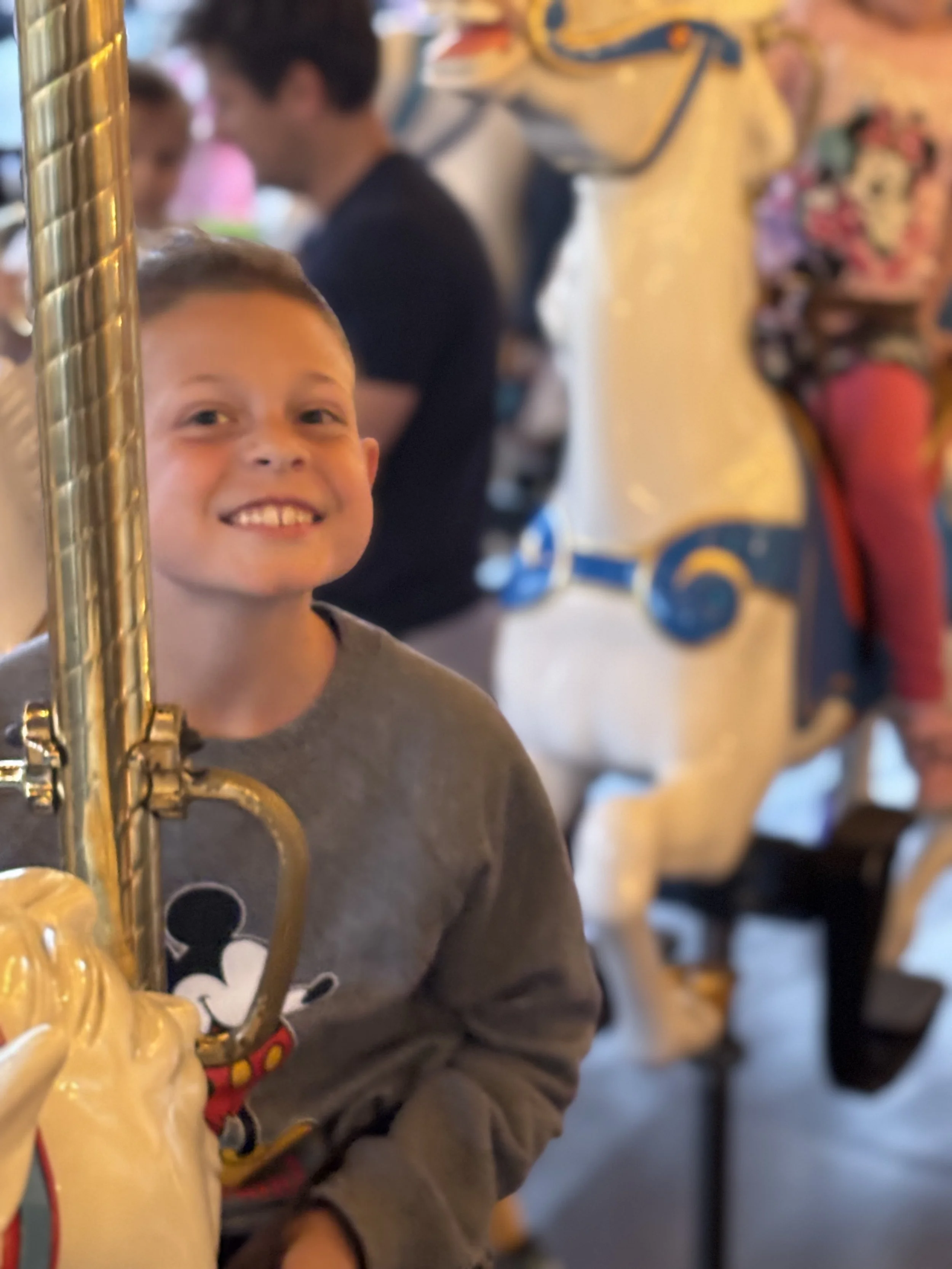 Close-up of a boy with a big smile holding a yellow saxophone in a busy, colorful indoor setting with people and artwork in the background.