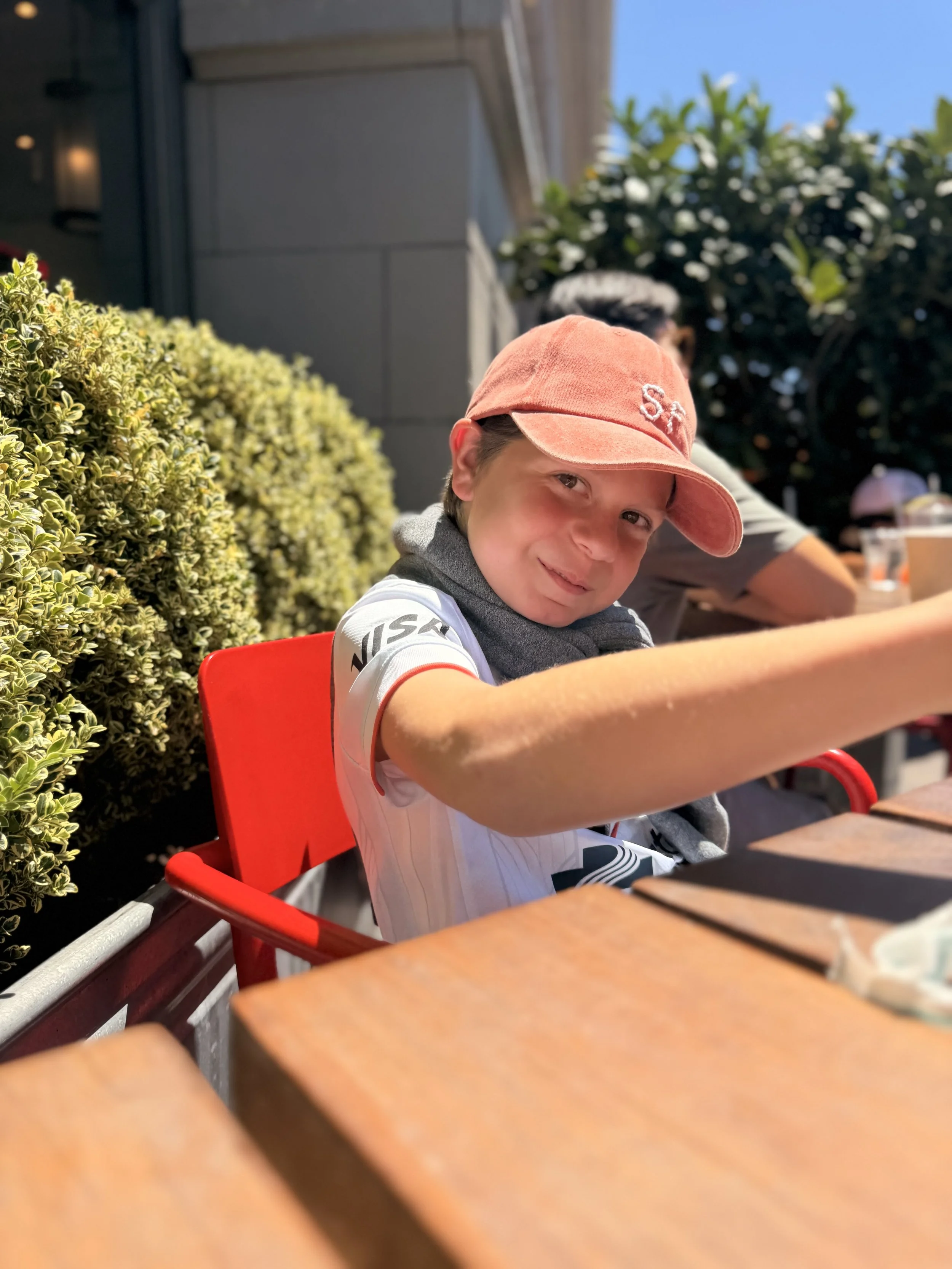 A young boy wearing a pink baseball cap and a white shirt with black accents, sitting outdoors at a table with a cheerful expression, surrounded by greenery and other people in the background.