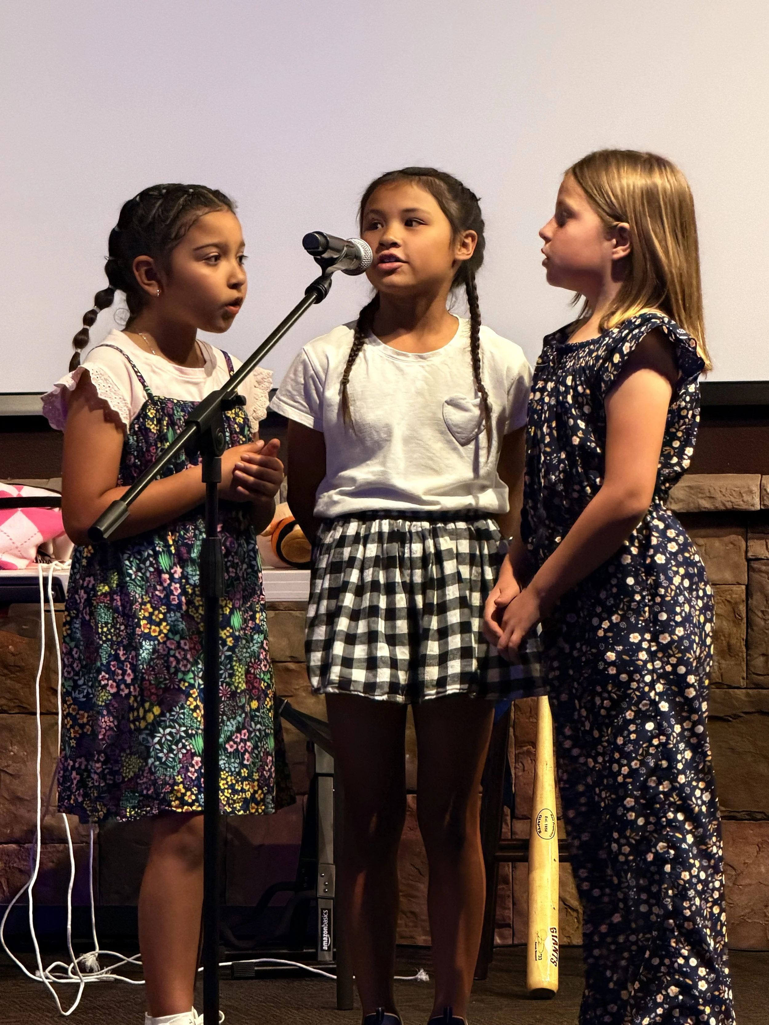 Three young girls standing and speaking or singing into a microphone, with a plain wall background and a yellow baseball bat on the floor behind them.