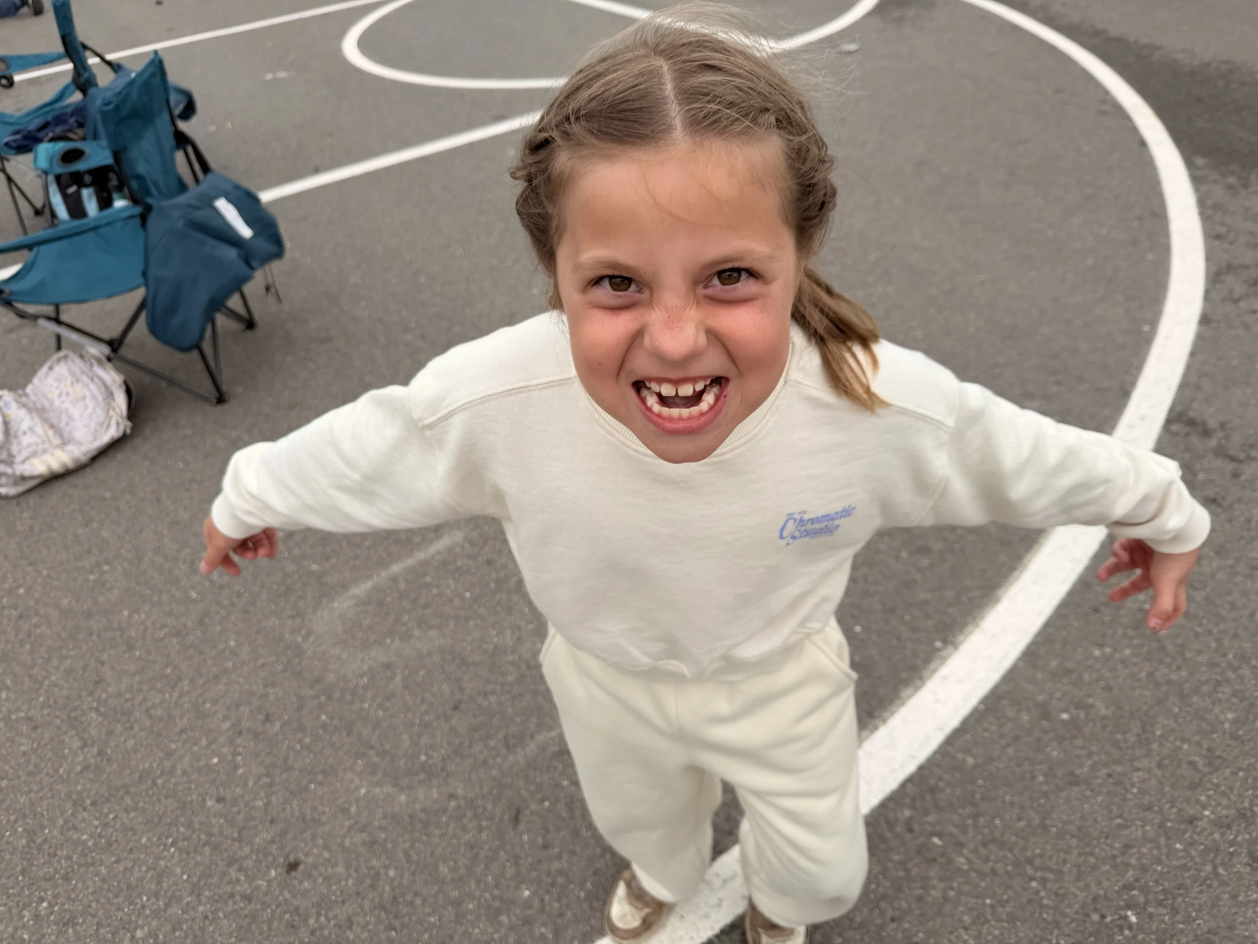 A young girl with light brown hair in braids, smiling widely with her mouth open, wearing a cream-colored sweatshirt and khaki pants, standing on a basketball court with white lines and a blue stroller in the background.