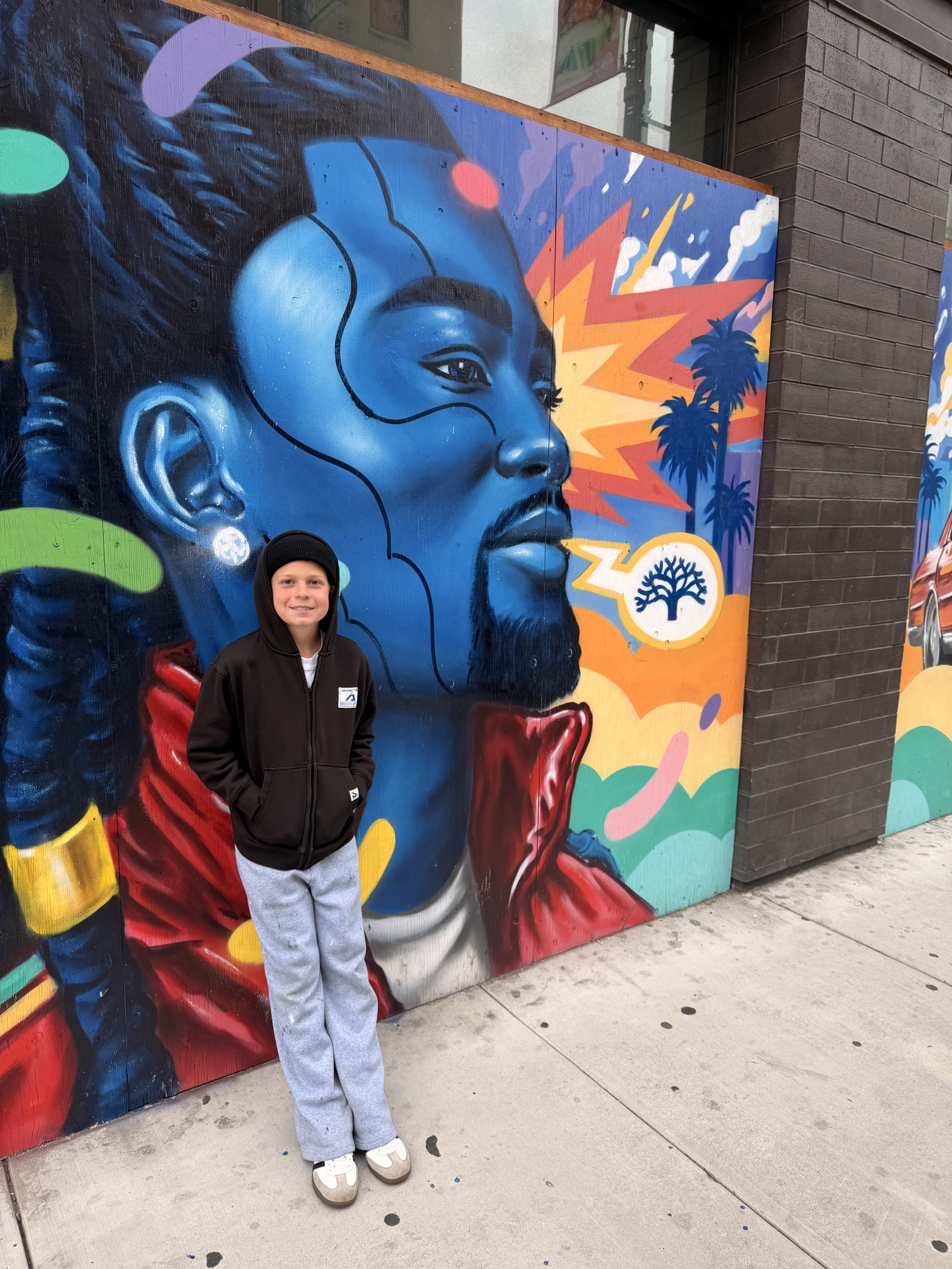 A young boy wearing a black hoodie and gray sweatpants standing in front of a colorful mural featuring a portrait of a woman with blue shading and red clothing, set against vibrant orange, purple, and yellow abstract background with palm trees.