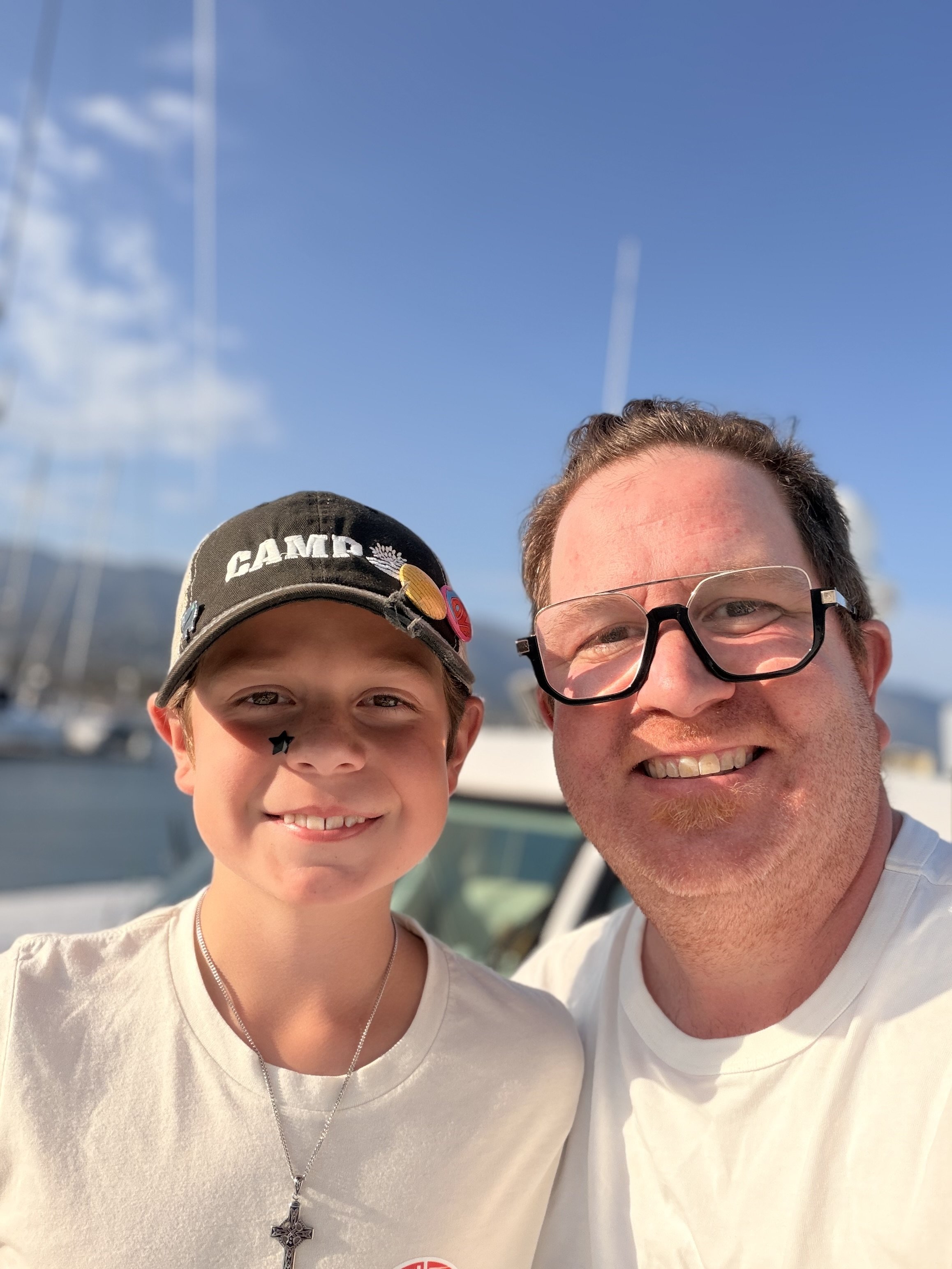 A young boy and an adult man smiling for a photo outdoors near boats under a blue sky.