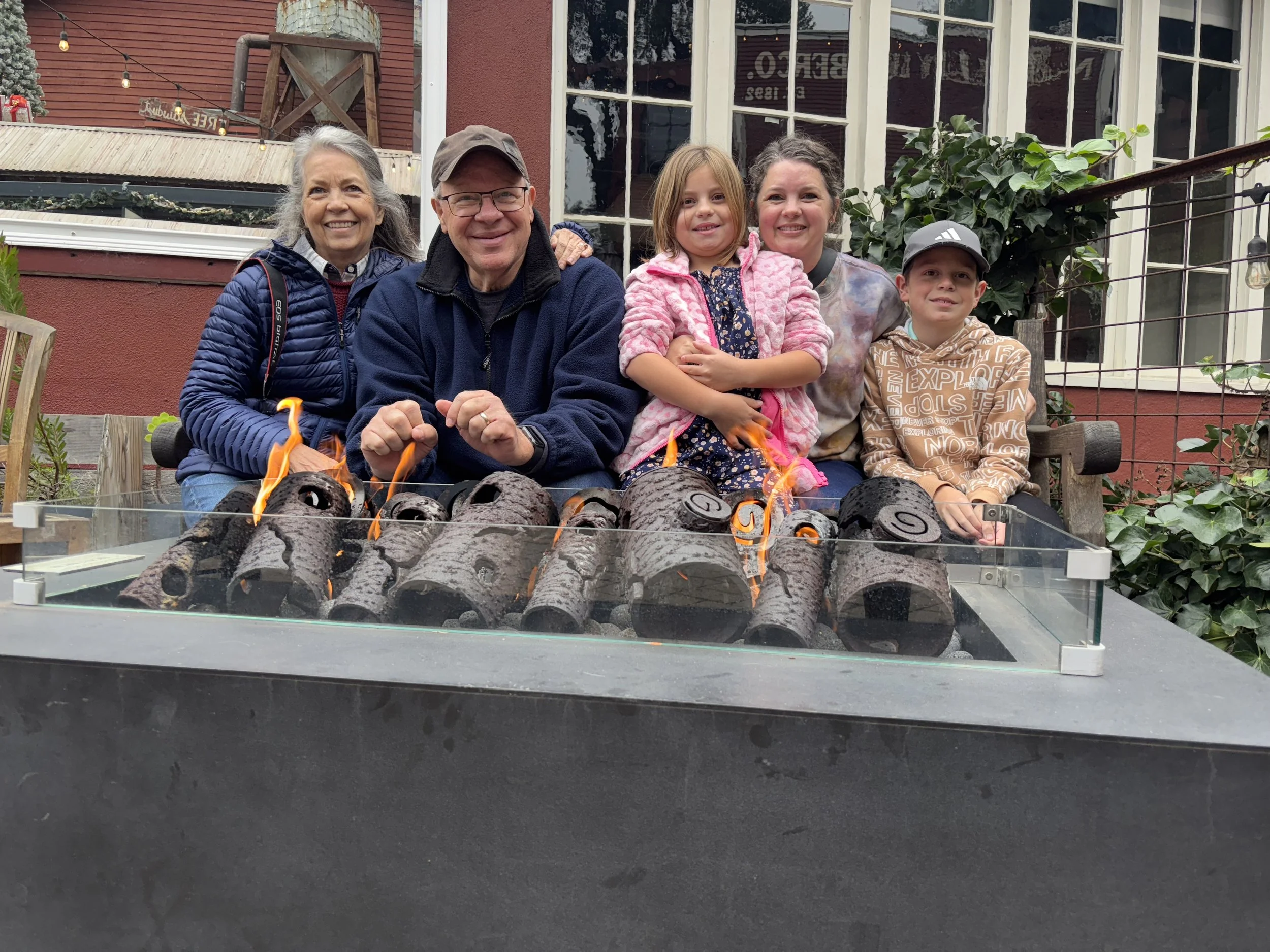 A group of five people, including two elderly adults and three children, sitting outdoors in front of a red house with large windows, next to a small fire pit with burning logs. They are smiling and dressed in casual clothing, with a festive atmosphe