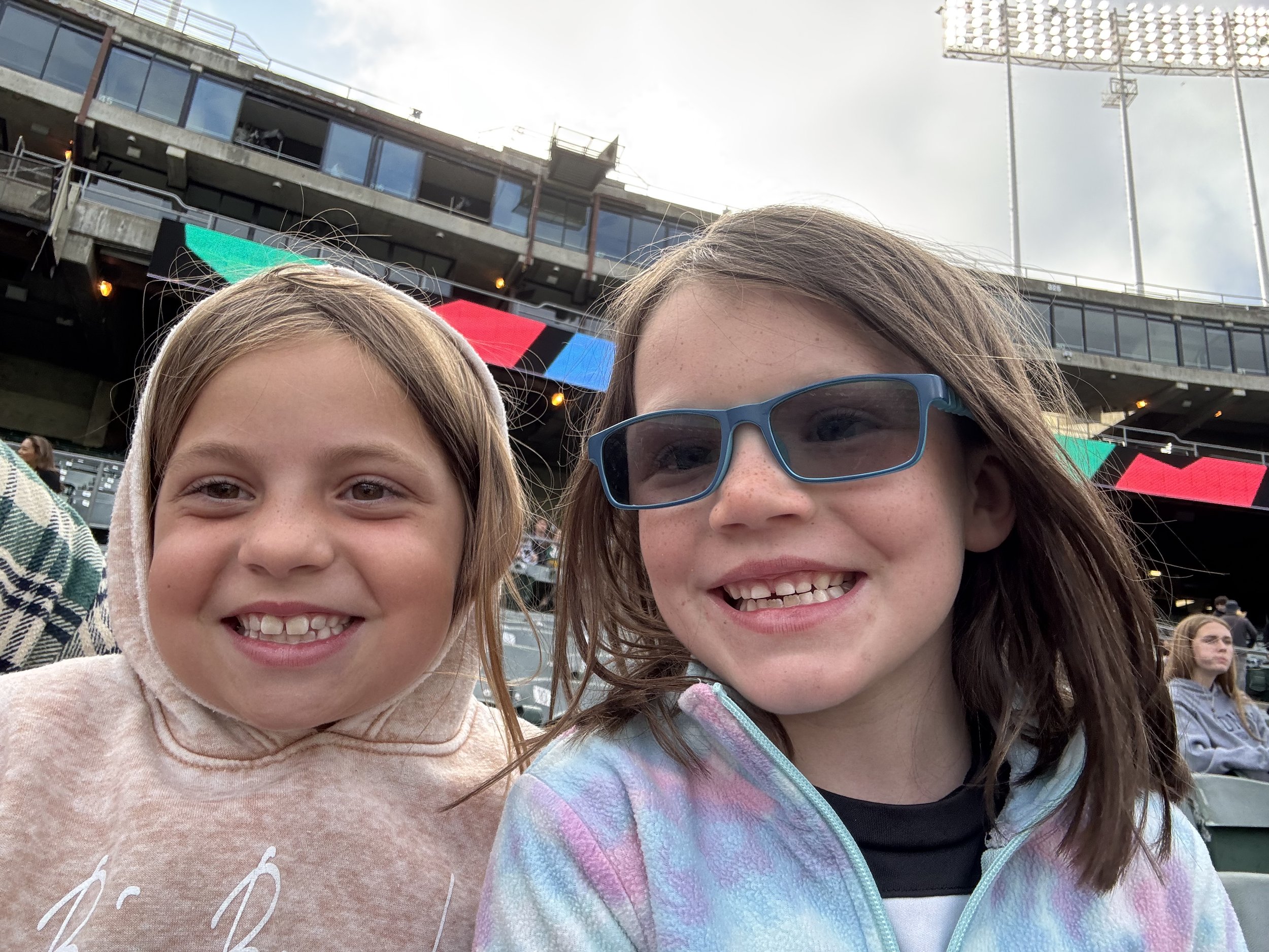 Two young girls smiling at an outdoor event with a cloudy sky and stage lights in the background.