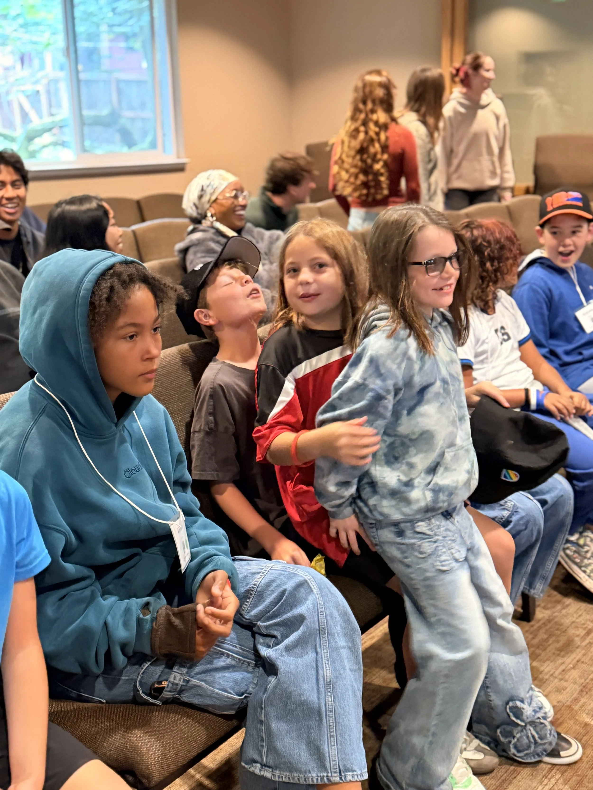 Children sitting and standing in a room, some smiling and engaging with each other.