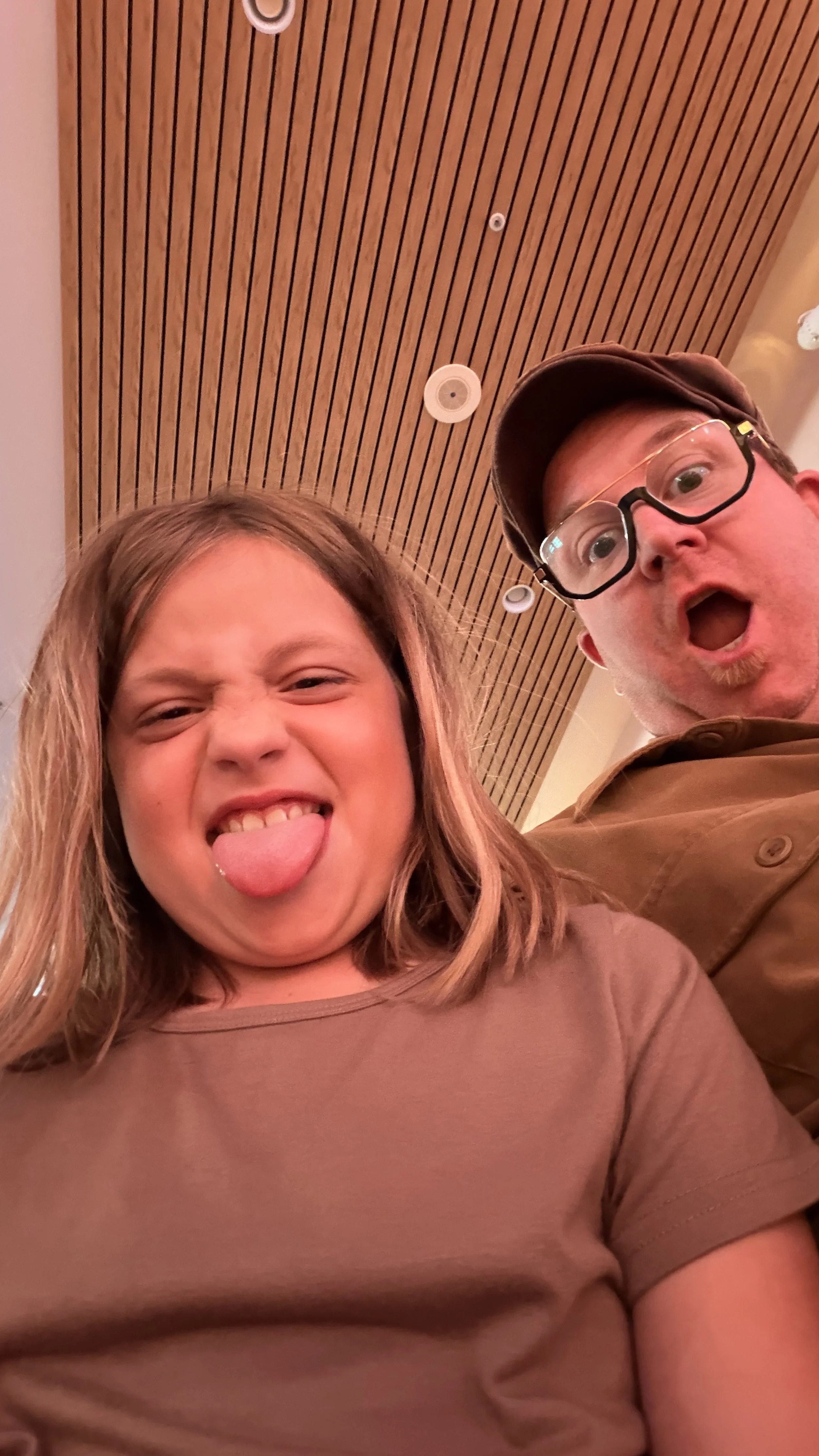 A young girl with light brown hair making a funny face by sticking out her tongue, and a man with glasses and a cap behind her with his mouth open in surprise or excitement, both in an indoor setting with a wooden ceiling.