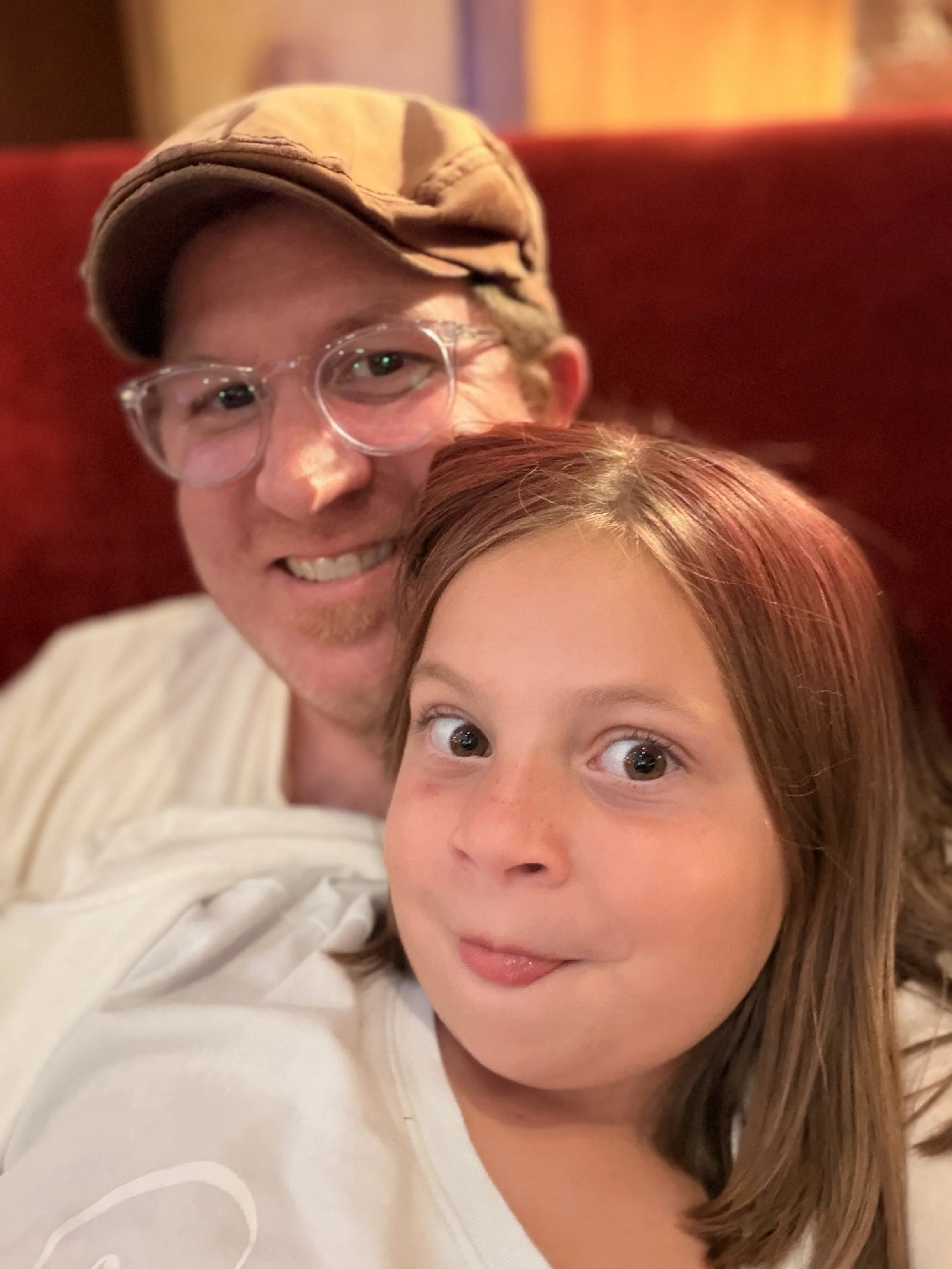 A man wearing clear glasses and a tan cap smiling, with a young girl with straight, light brown hair and big eyes in front of him. They are close together indoors.