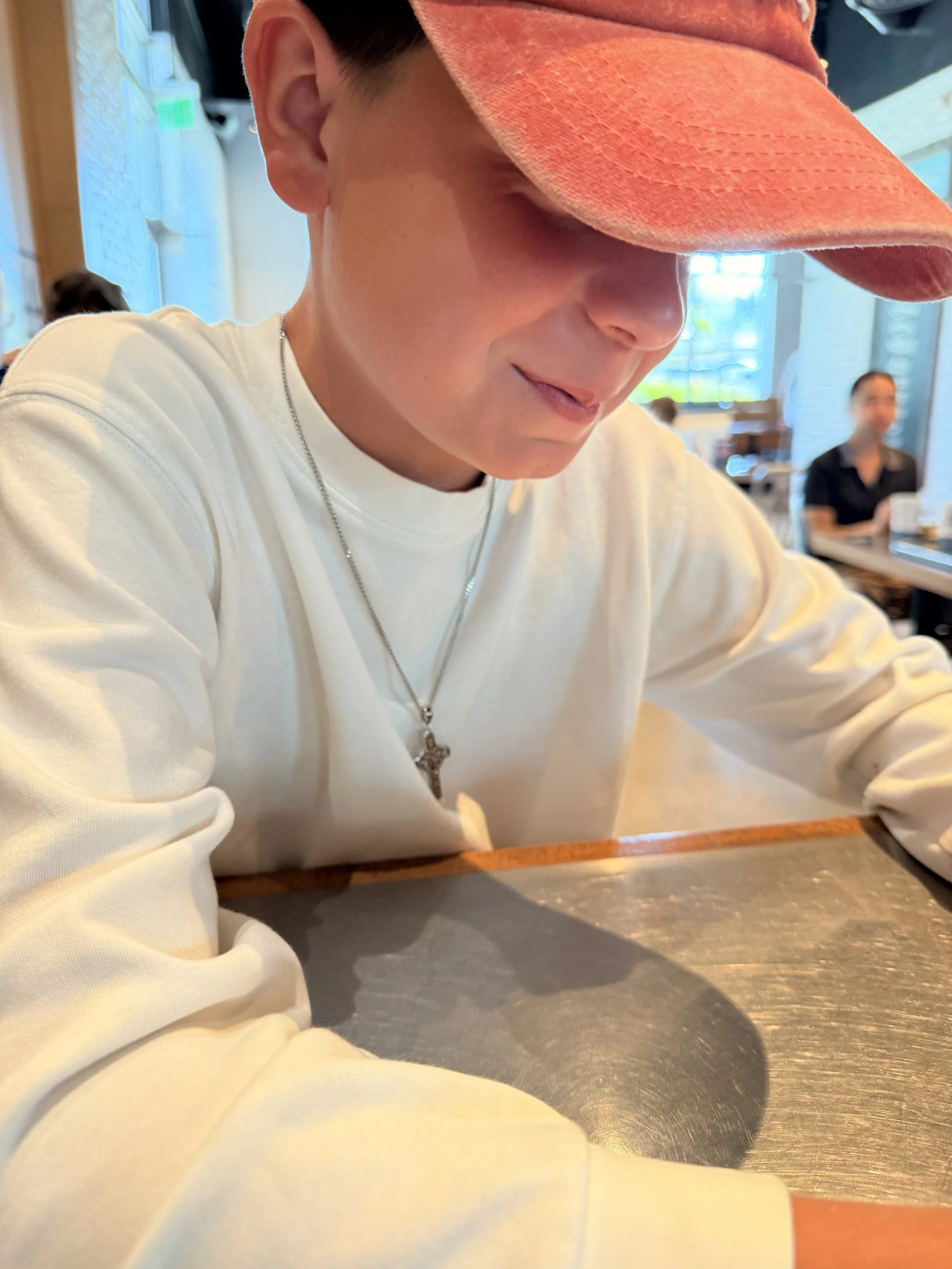 Close-up of a person wearing an orange cap and a white sweatshirt, sitting at a table in a restaurant with a background of large windows and other diners.