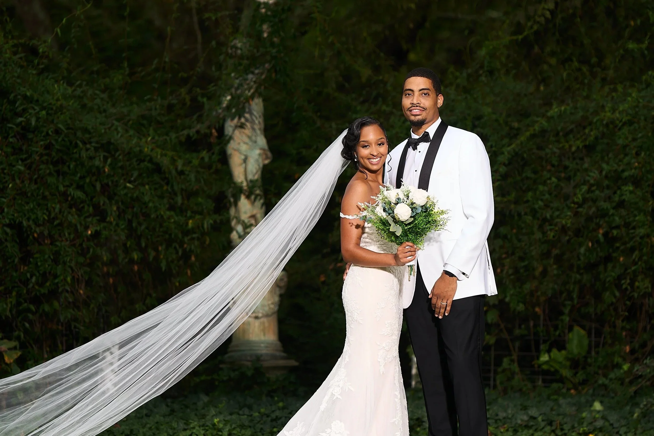 A newlywed couple standing outdoors in front of greenery, with the bride holding a bouquet and wearing a white lace wedding gown with a veil, and the groom in a white tuxedo jacket with black lapels and a black bow tie.